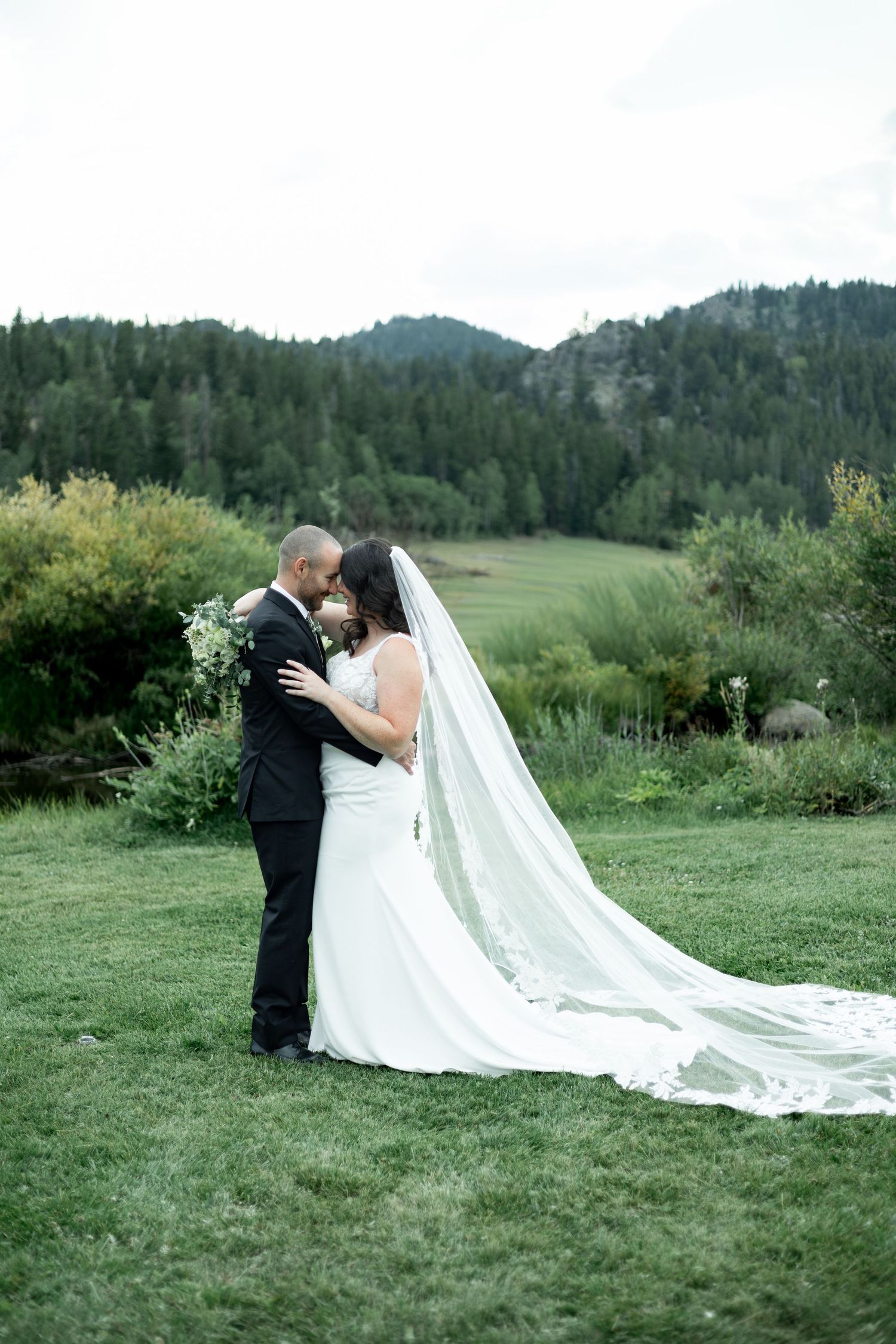 Bride and groom share a romantic moment on lush green lawn with mountain forest backdrop on their wedding day.
