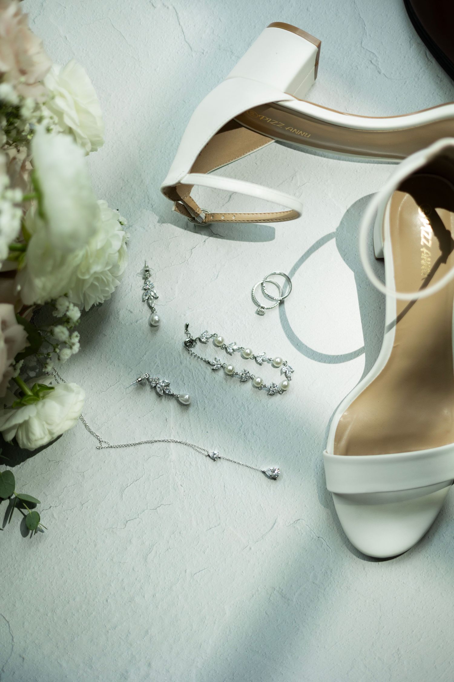 Elegant white bridal shoes and delicate floral accessories arranged on a light textured surface.