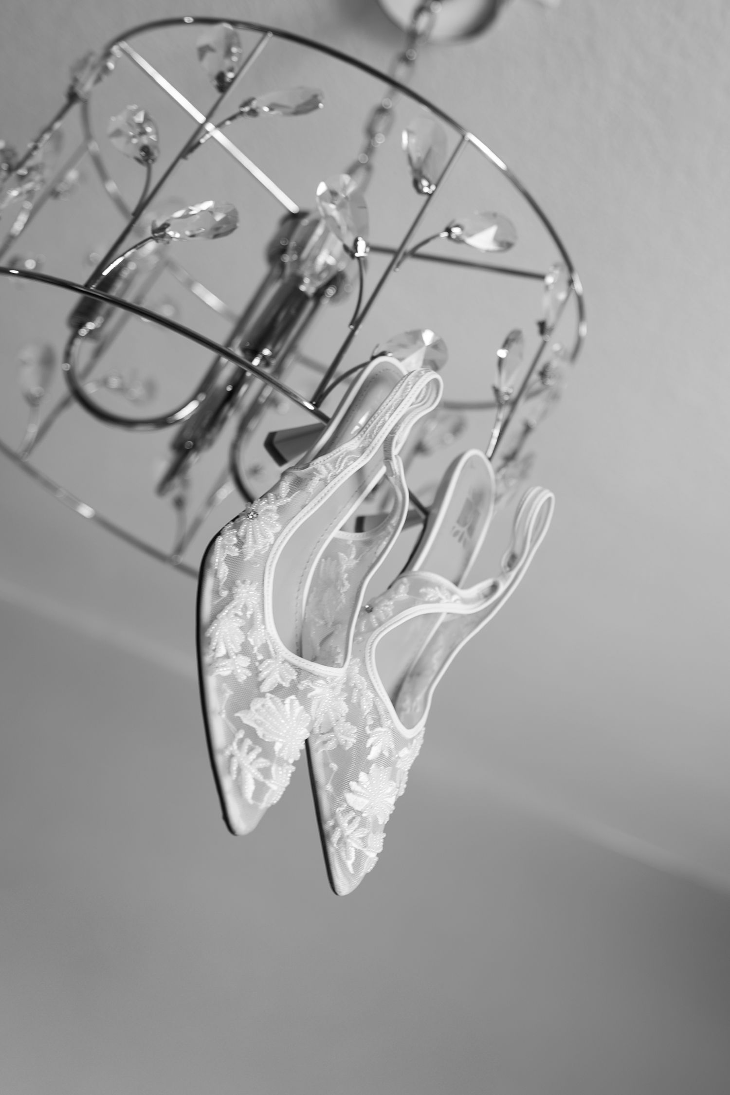 Delicate lace wedding shoes hanging from a crystal chandelier in black and white photography.