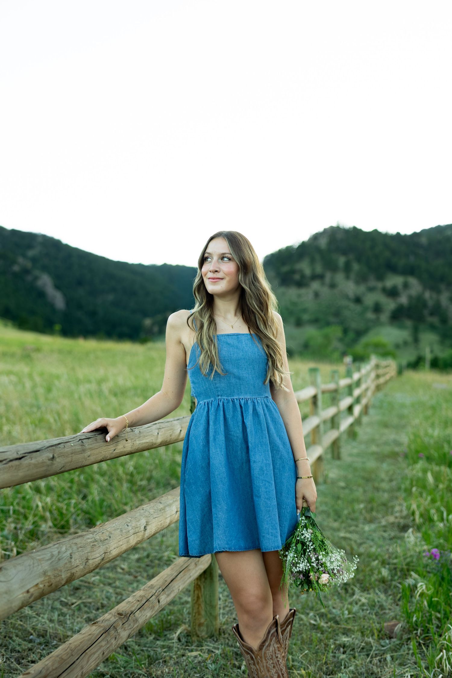 Young woman in blue denim dress and cowboy boots holding wildflowers by a wooden fence in a green mountain meadow.