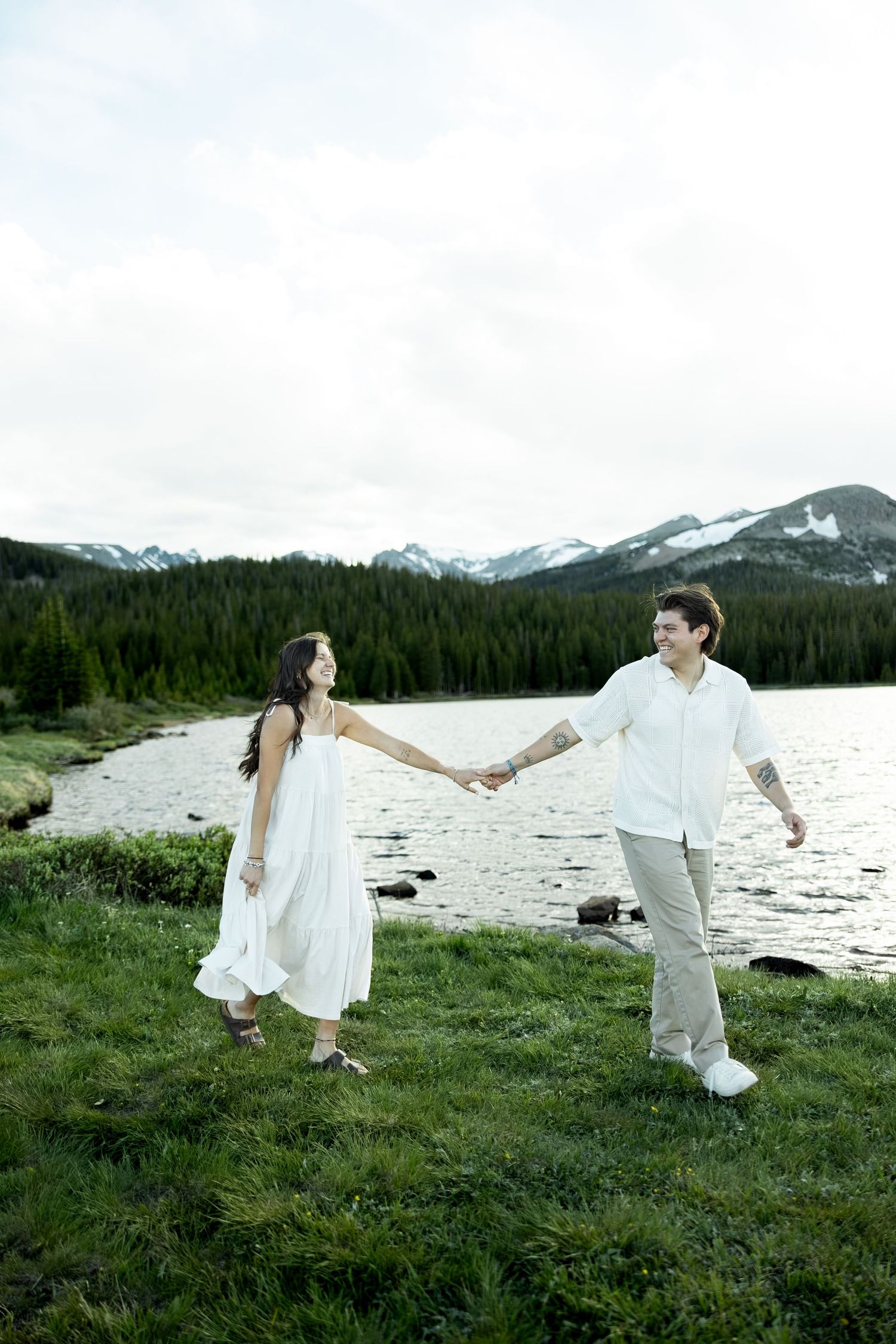 Couple holding hands and walking by a mountain lake, woman in white dress, man in white shirt, with snowy peaks behind.