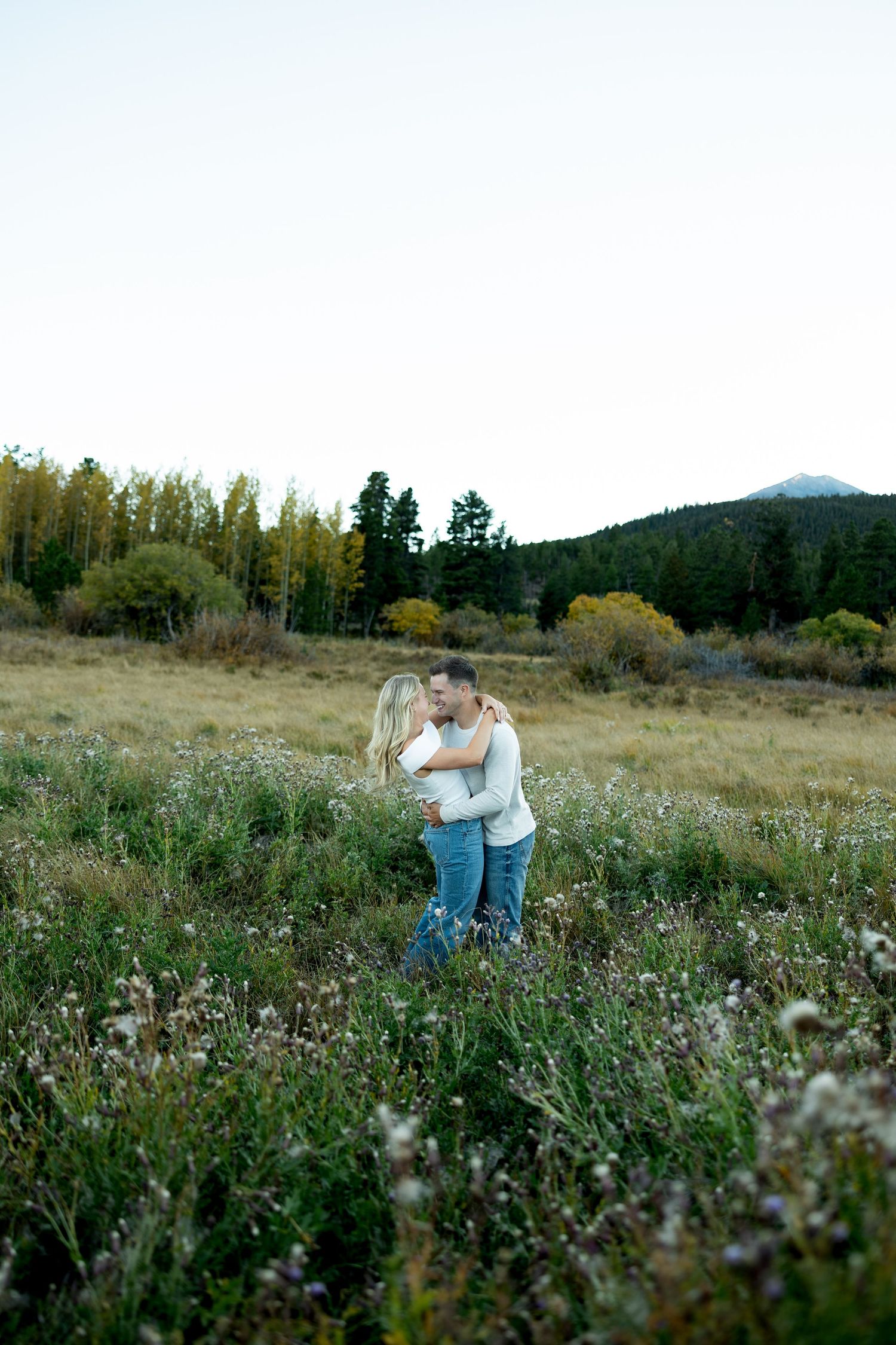 Couple embracing in a wildflower meadow with golden aspen trees and mountains in the background during an outdoor engagement photo session.