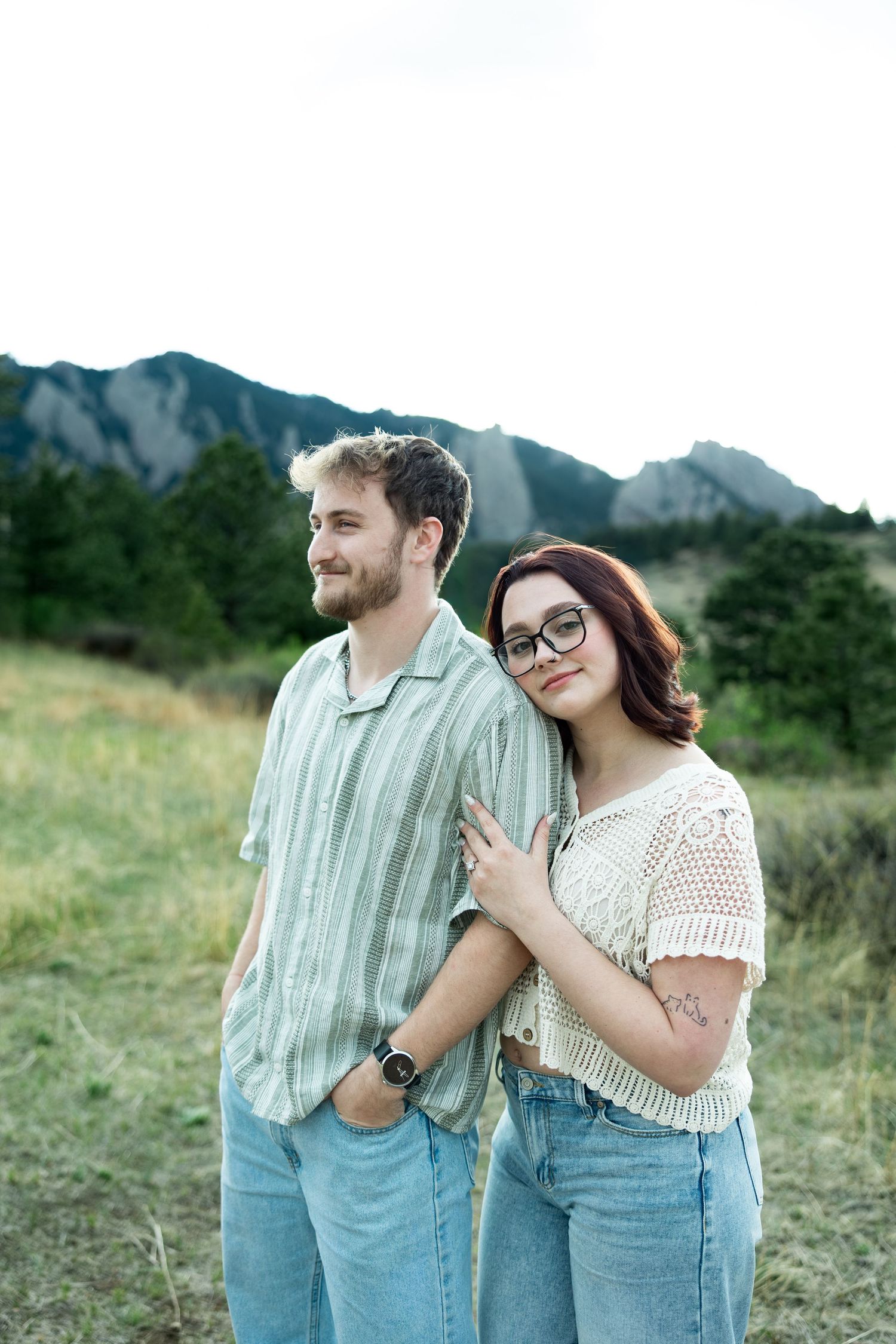 A couple poses in a mountain meadow, woman in crochet top leaning on man in striped shirt, both wearing jeans.