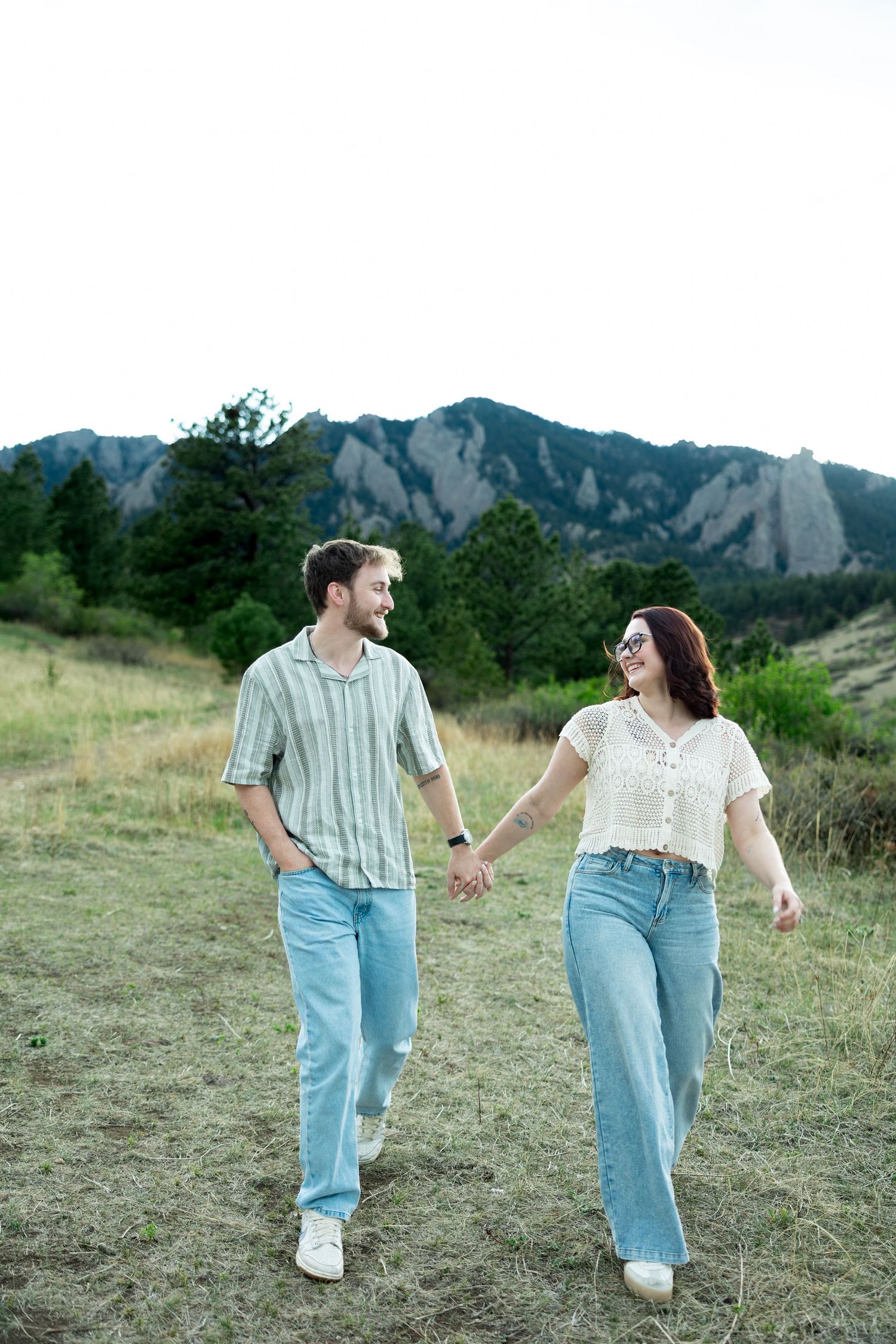 Couple holding hands walking through mountain meadow, smiling at each other, wearing casual jeans outfits with rocky peaks behind.