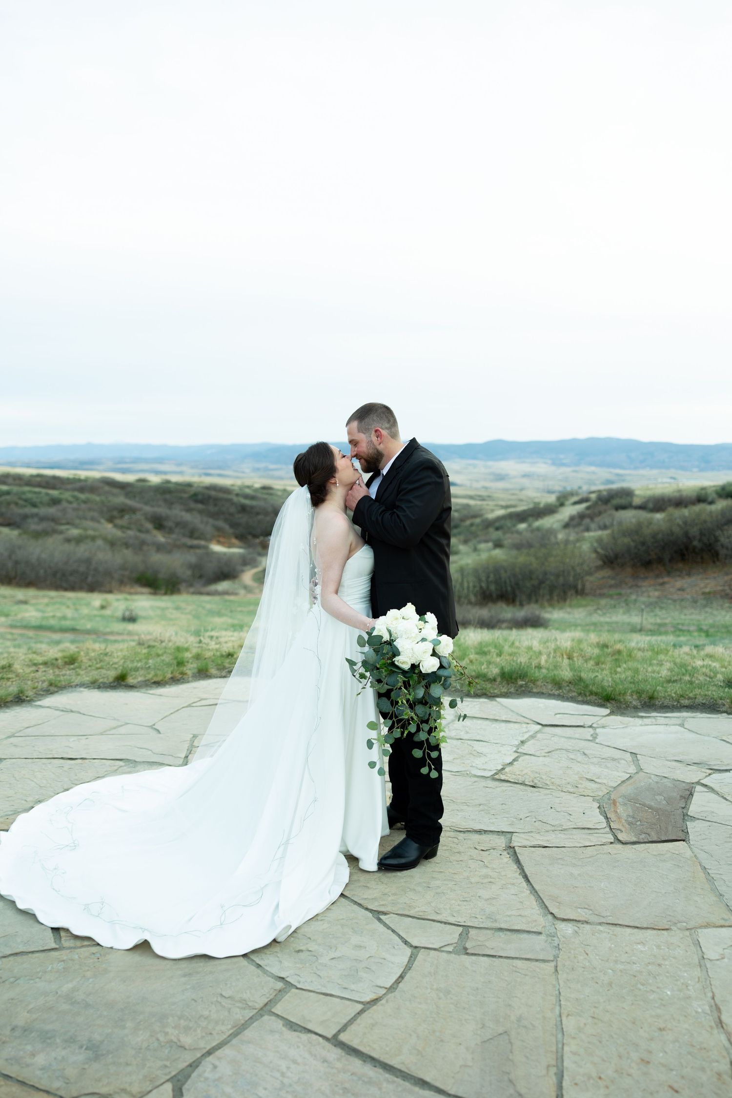 Bride and groom share an intimate moment on a stone terrace with sweeping countryside views, bride holding white bouquet.