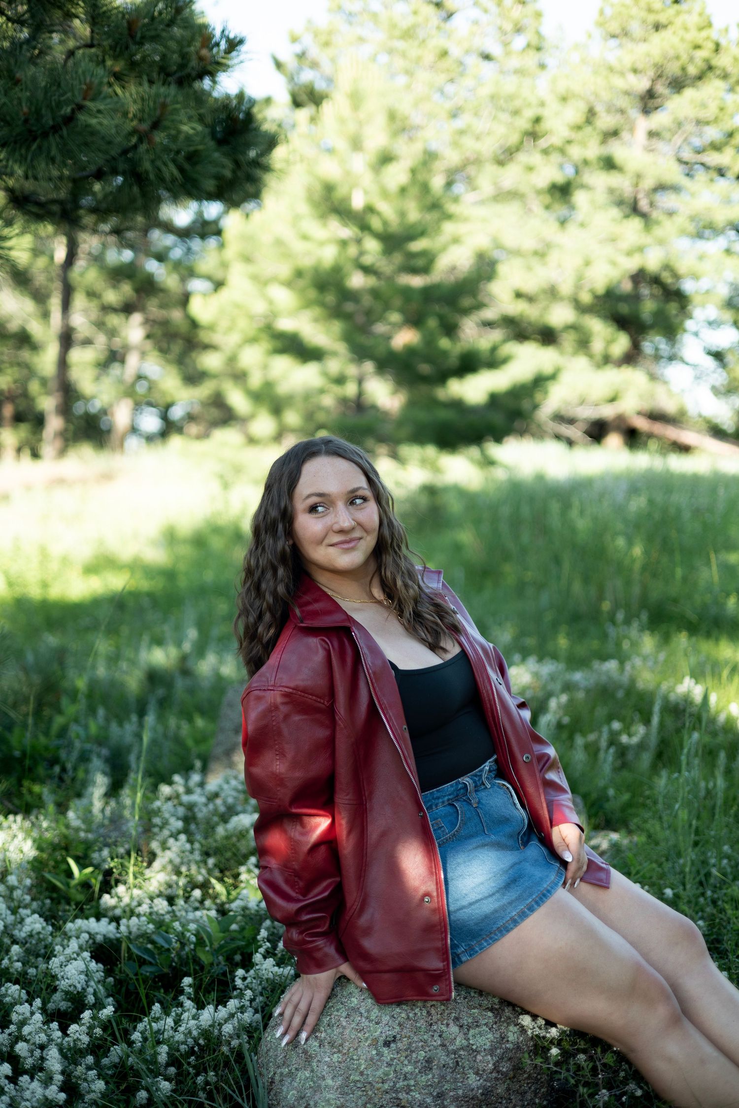 A person in a red leather jacket and denim shorts sits on a rock in a sunny outdoor setting with pine trees.