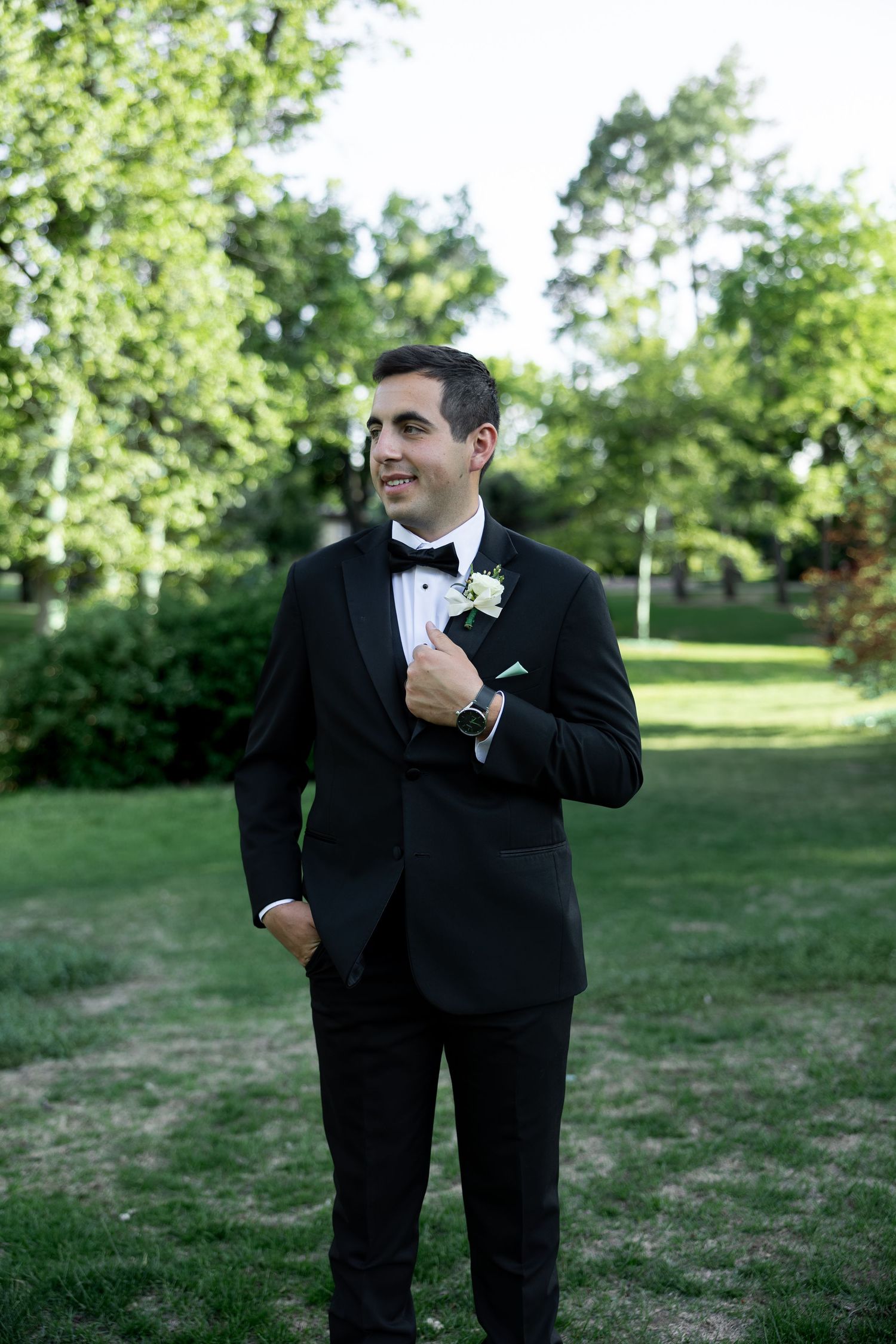 Handsome groom in black tuxedo with boutonniere stands outdoors in lush green park setting.