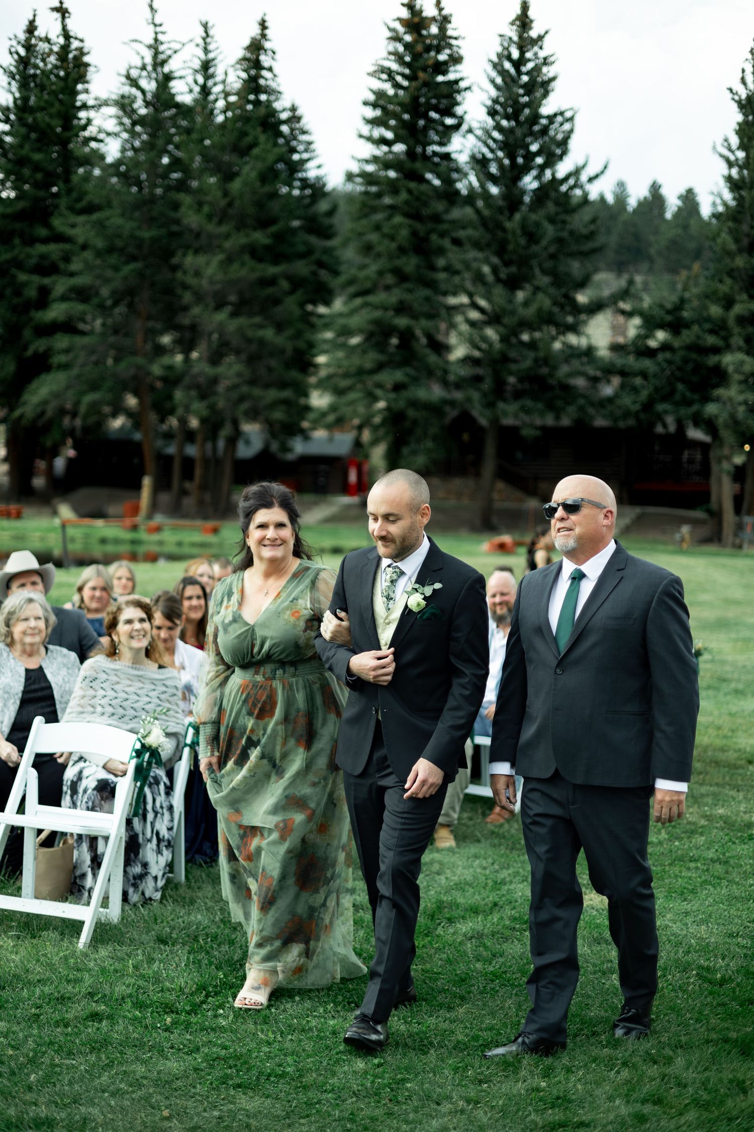 Wedding guests mingle on a grassy lawn surrounded by tall evergreen trees during an outdoor ceremony.