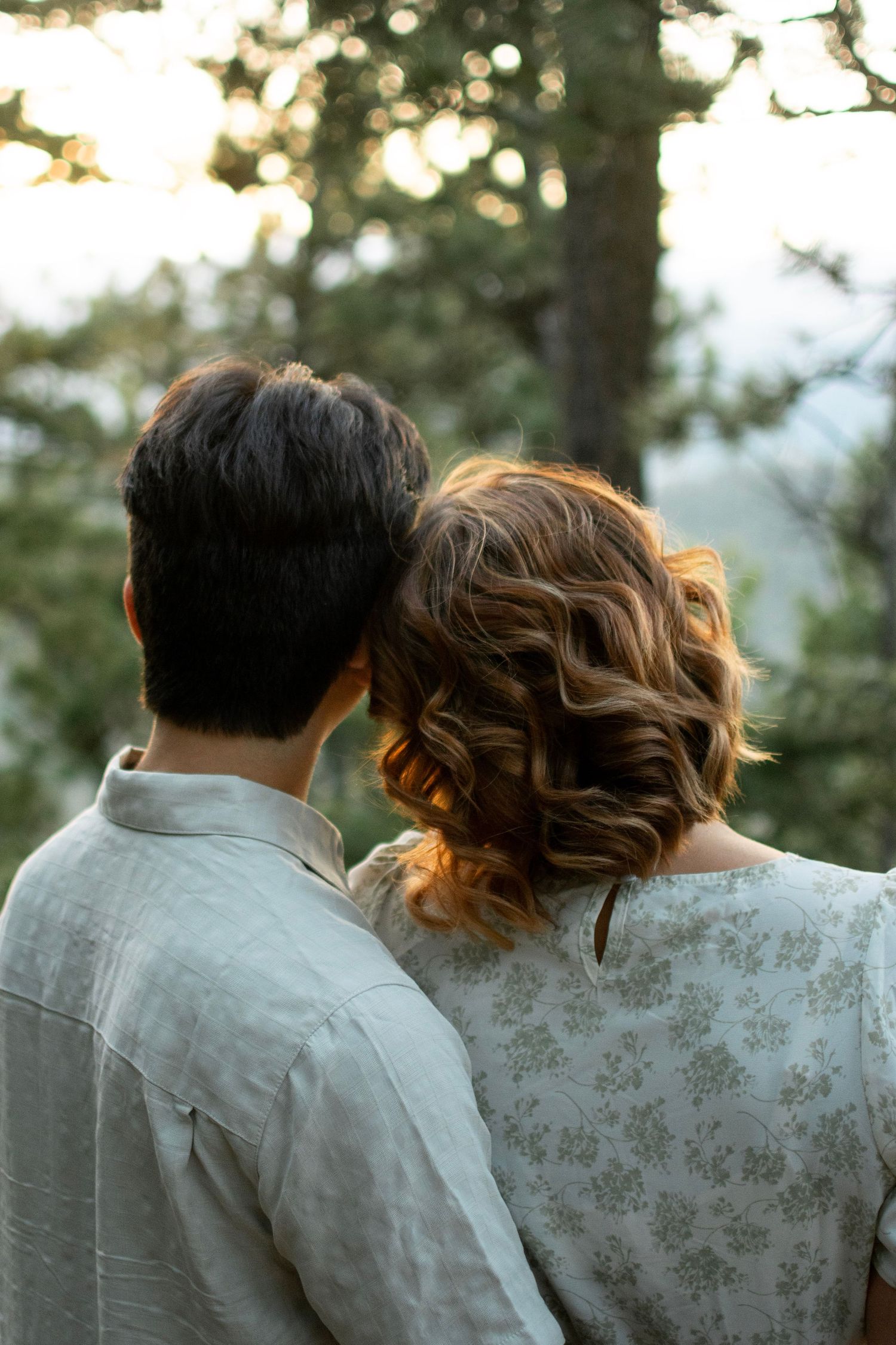Couple in light colored clothing share an intimate moment against a sunlit forest backdrop.