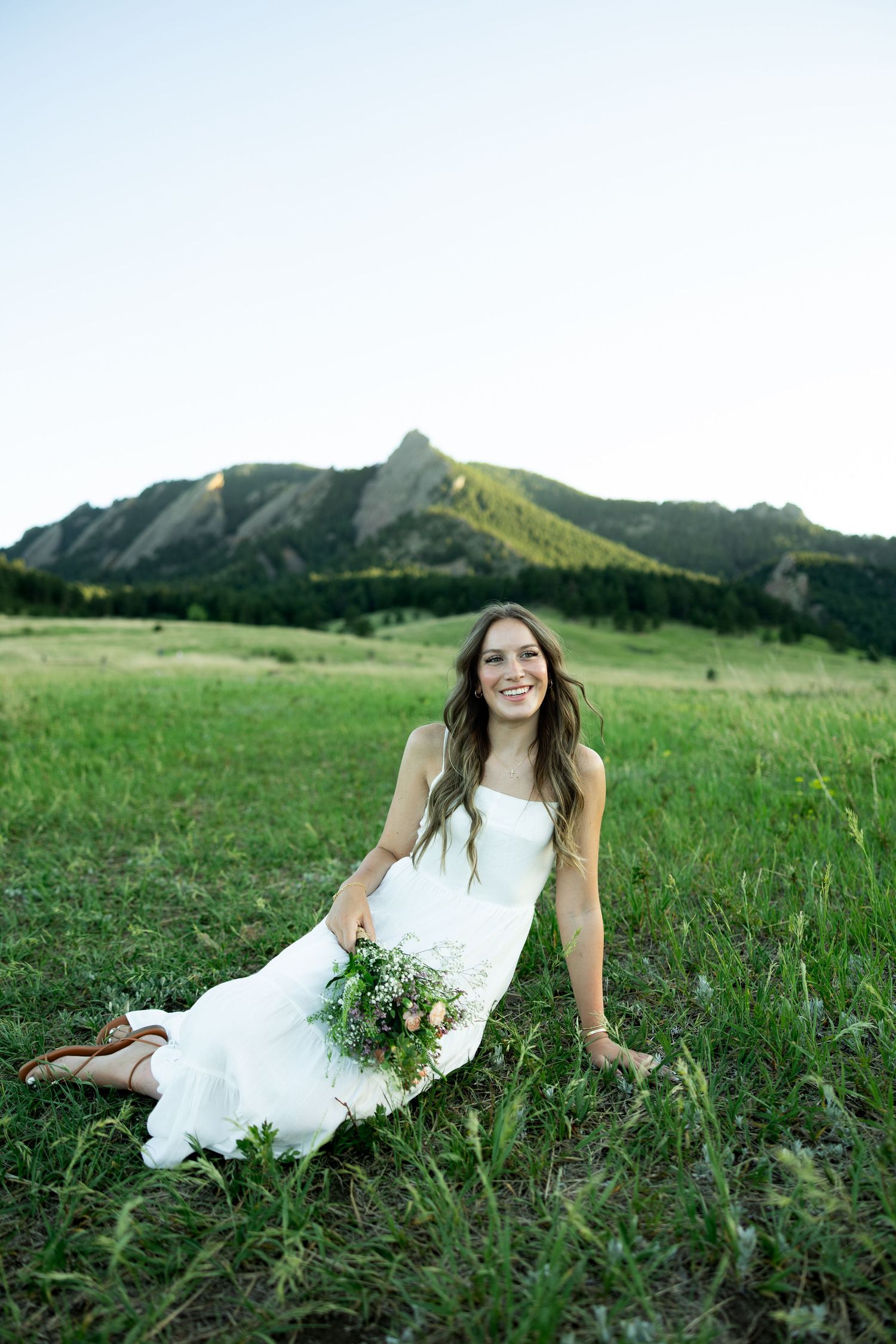 Smiling bride in white dress sitting in green meadow with bouquet, rocky mountain flatirons in background.