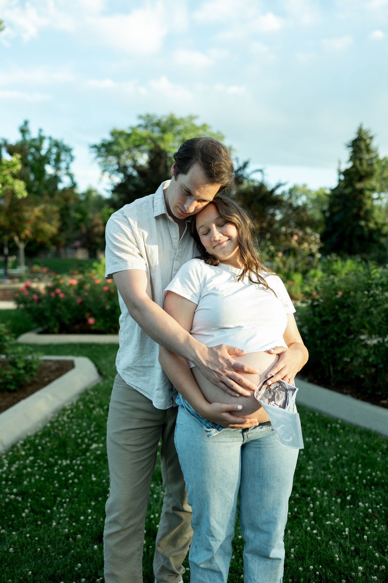 Loving couple embracing in a park, man hugging pregnant woman from behind while holding ultrasound photos.