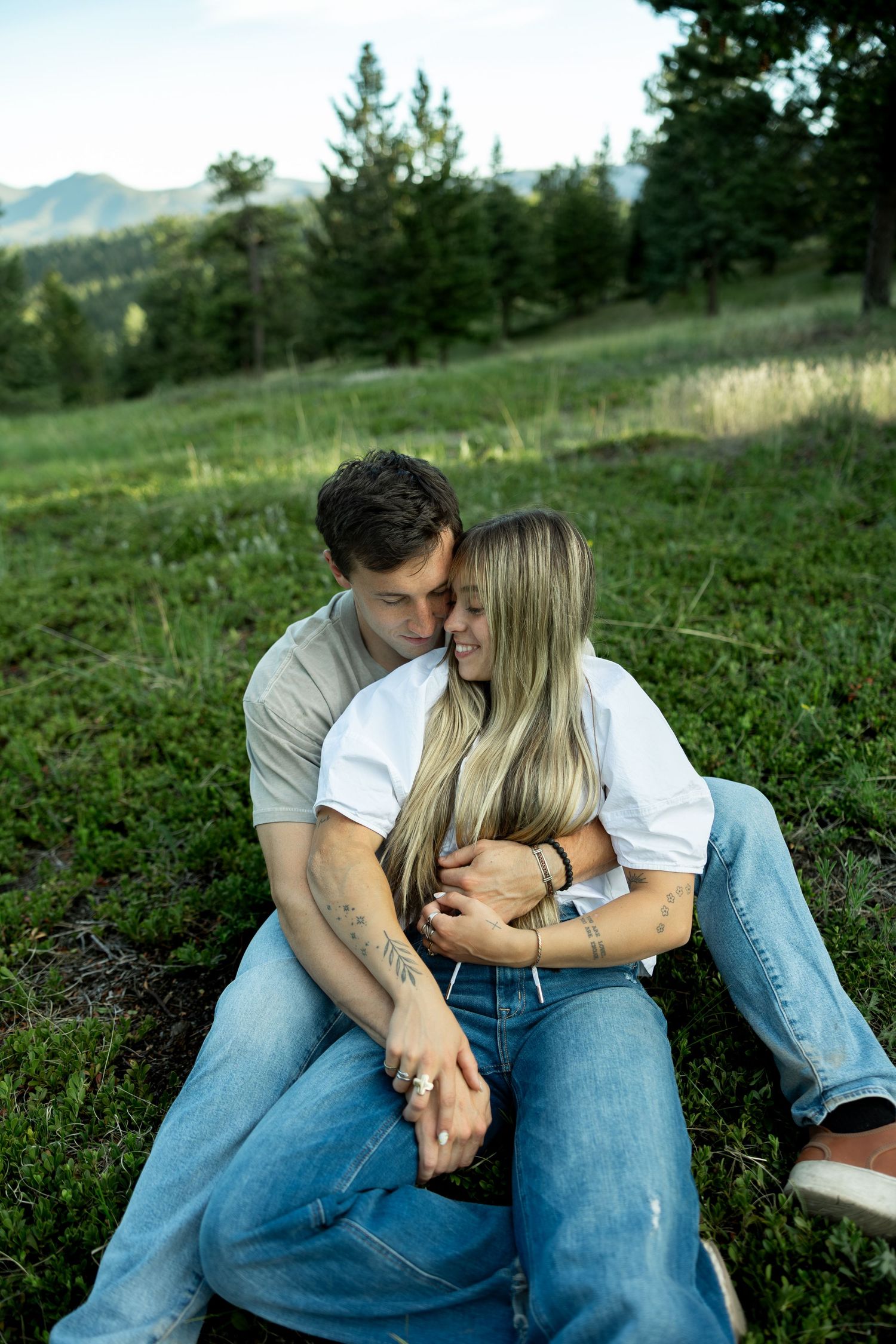 A couple sits together on a grassy hillside, embracing tenderly with mountains and pine trees in the background.