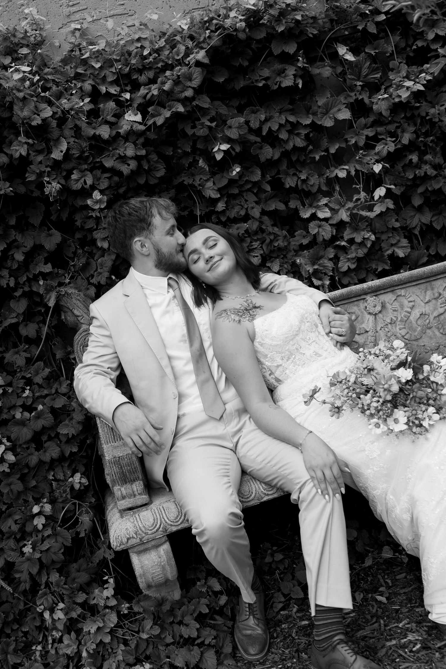 Bride and groom share a romantic moment on a bench surrounded by ivy in black and white photo.