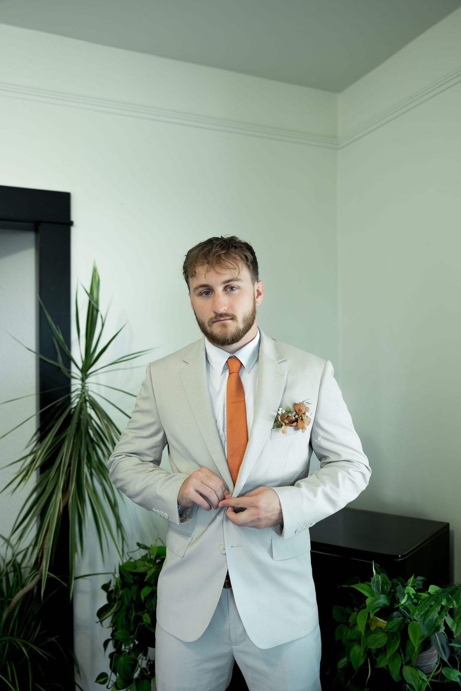 A groom in a light gray suit with an orange tie and boutonniere buttons his jacket while standing near green plants.