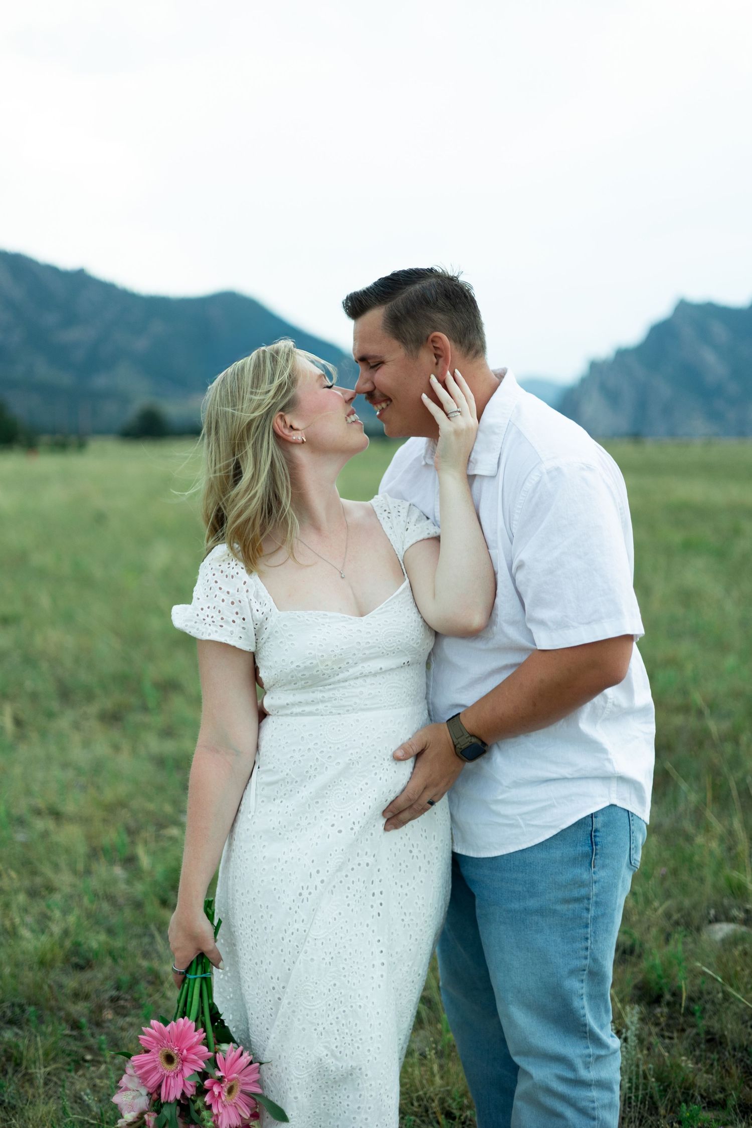 Couple in white outfits sharing a romantic moment in a green field with mountain views, woman holding pink flowers.