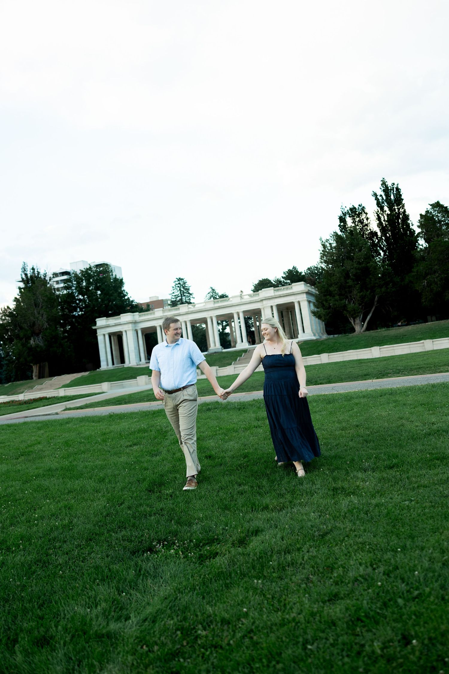 A couple holds hands while walking on green grass with a white columned pavilion and trees in the background.