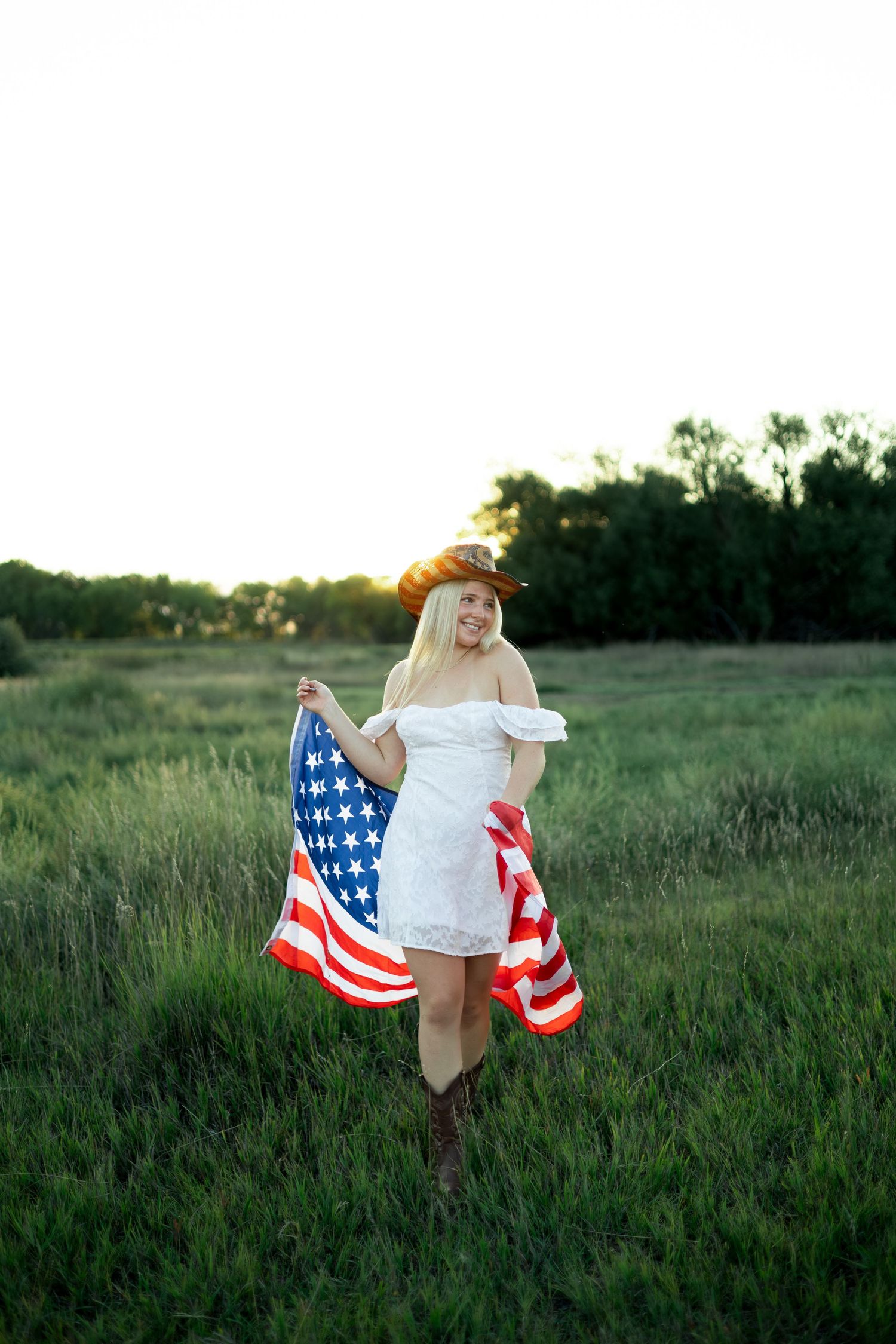 A person in a white dress holds an American flag while standing in a grassy field at sunset.
