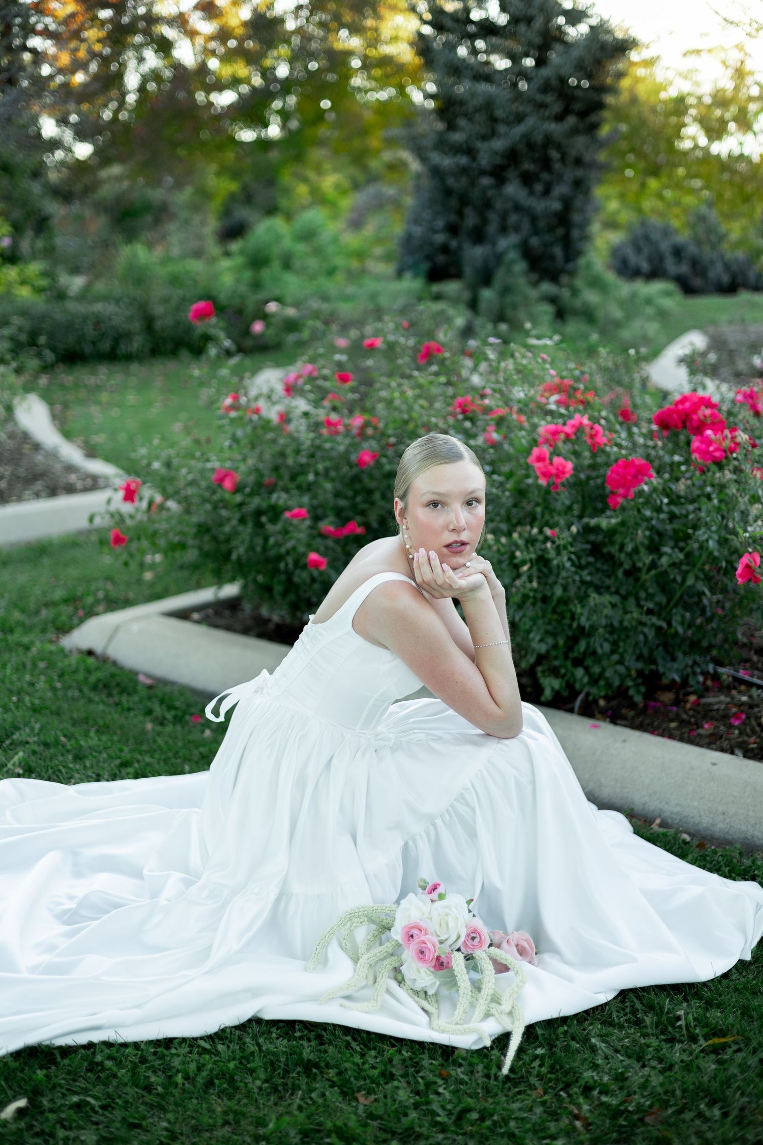 A white wedding dress flows across green grass near vibrant pink rose bushes in a garden setting.