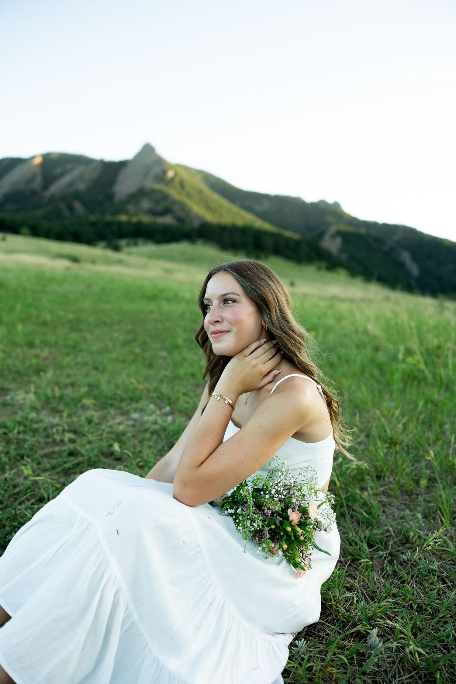 Young bride in white dress sits in green meadow holding wildflower bouquet with rocky mountain peaks in background.