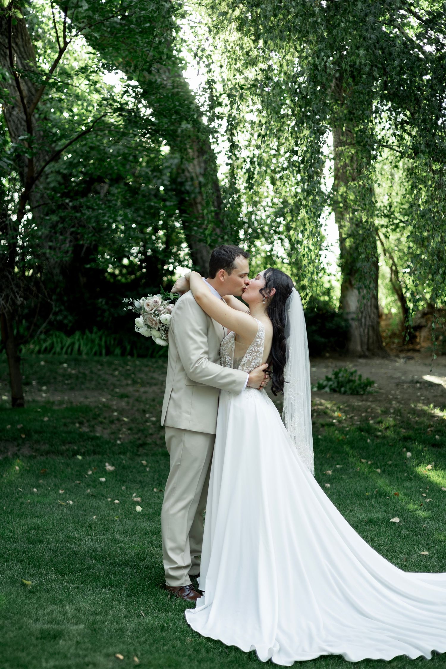 Bride and groom share a romantic embrace in a lush green garden, bride wearing a flowing white gown with veil.
