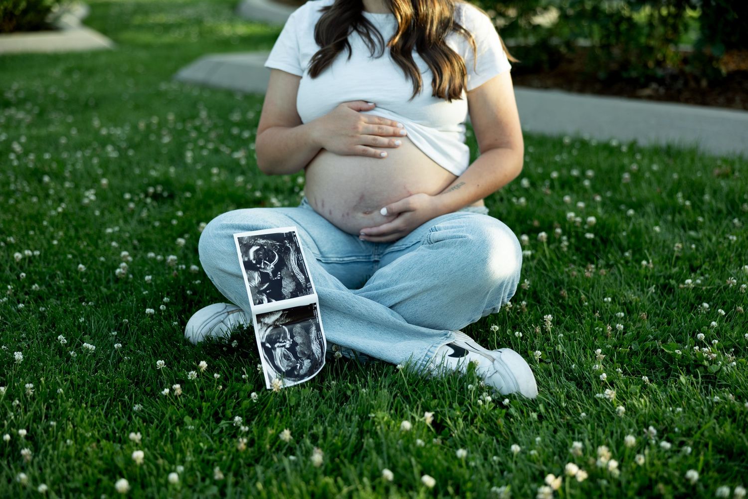 Pregnant woman sitting on grass holding baby bump with ultrasound photos, maternity outdoor photography.