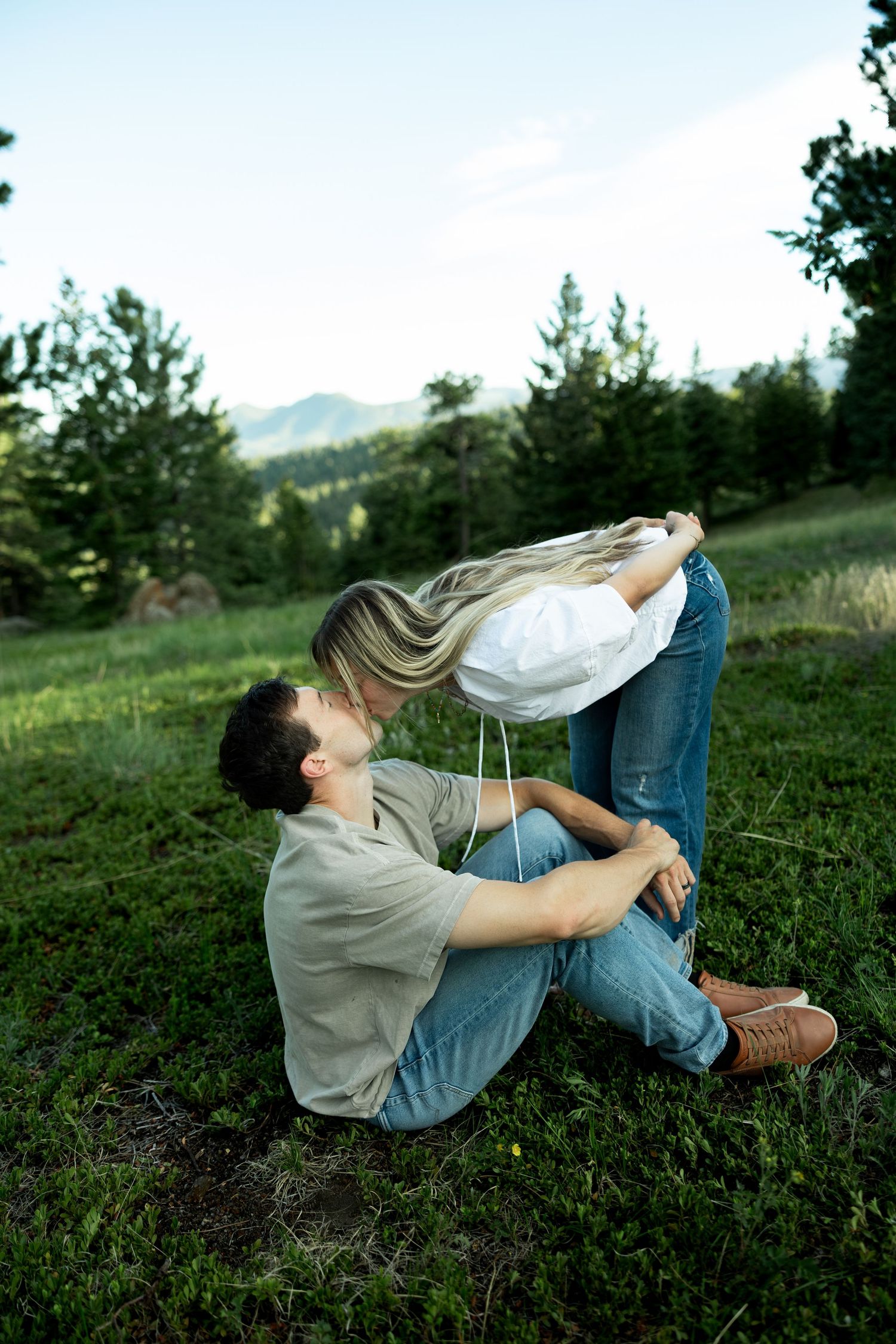 Couple sharing a romantic kiss in a lush green meadow surrounded by pine trees and mountains in the background.