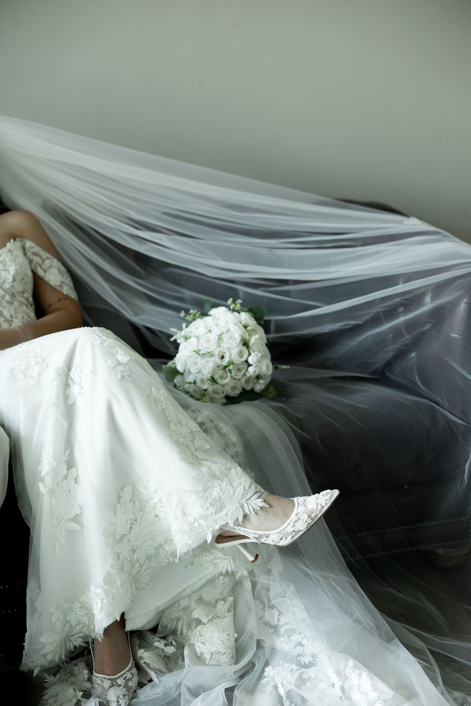 A white bridal bouquet rests on flowing tulle veil next to lace wedding dress and crystal shoes.