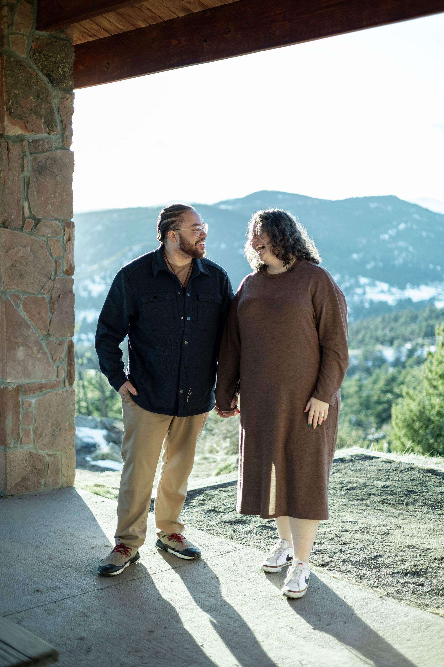Couple holding hands under a rustic stone pavilion with mountain views in the background.