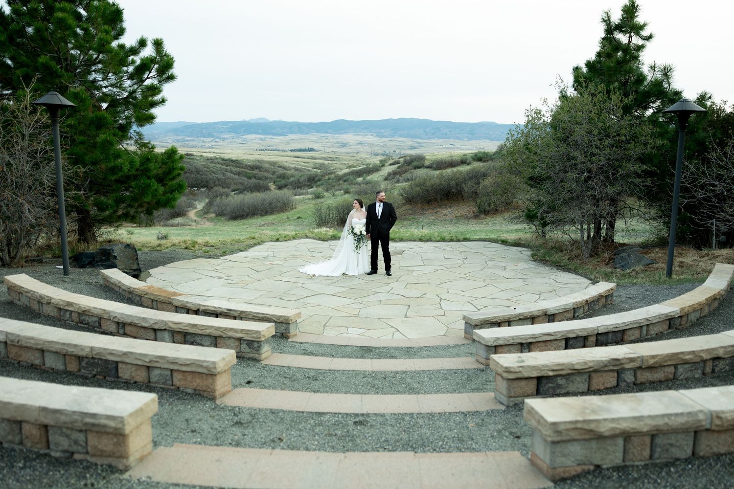 Bride and groom stand together at an outdoor stone ceremony venue overlooking a scenic valley landscape.