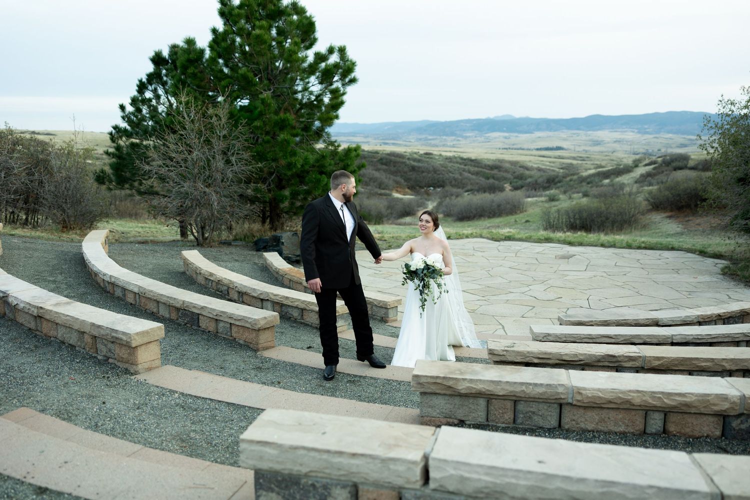 Bride and groom walk hand-in-hand through a stone amphitheater with scenic rolling hills backdrop.