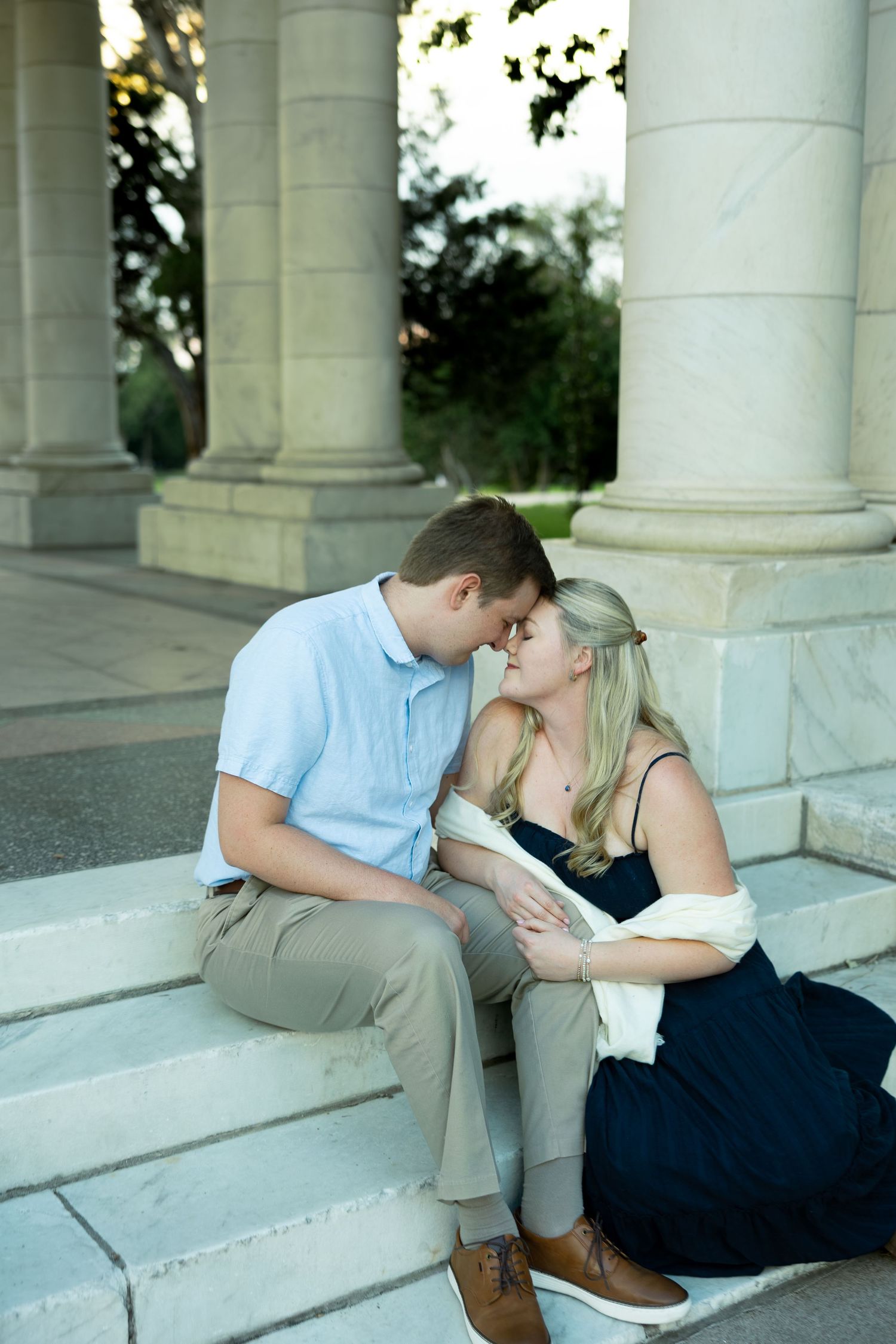 Couple sharing a romantic moment on marble steps between columns, woman in navy dress, man in light blue shirt.