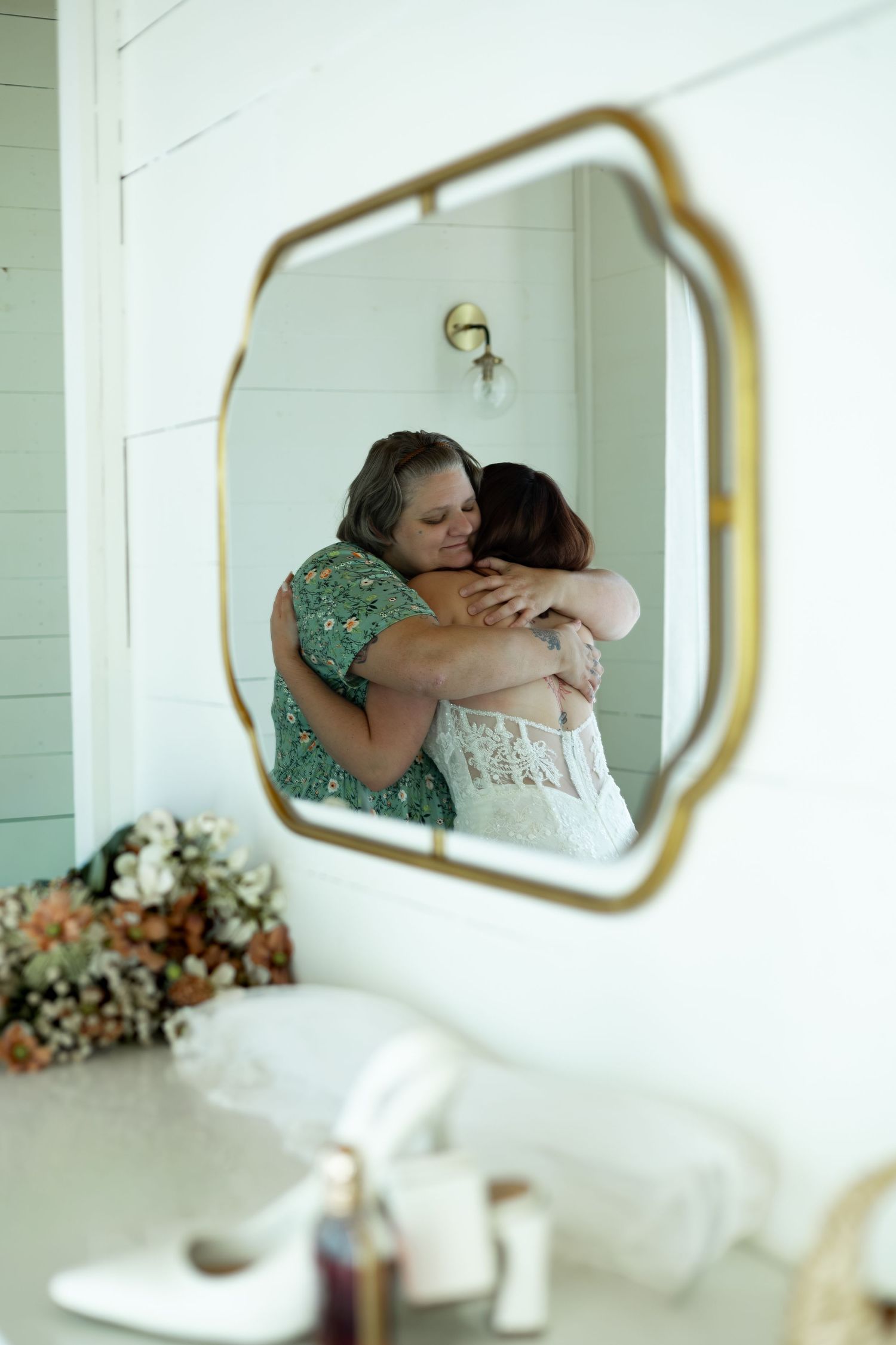 Two women share a heartfelt hug reflected in a gold ornate mirror during bridal wedding preparations.