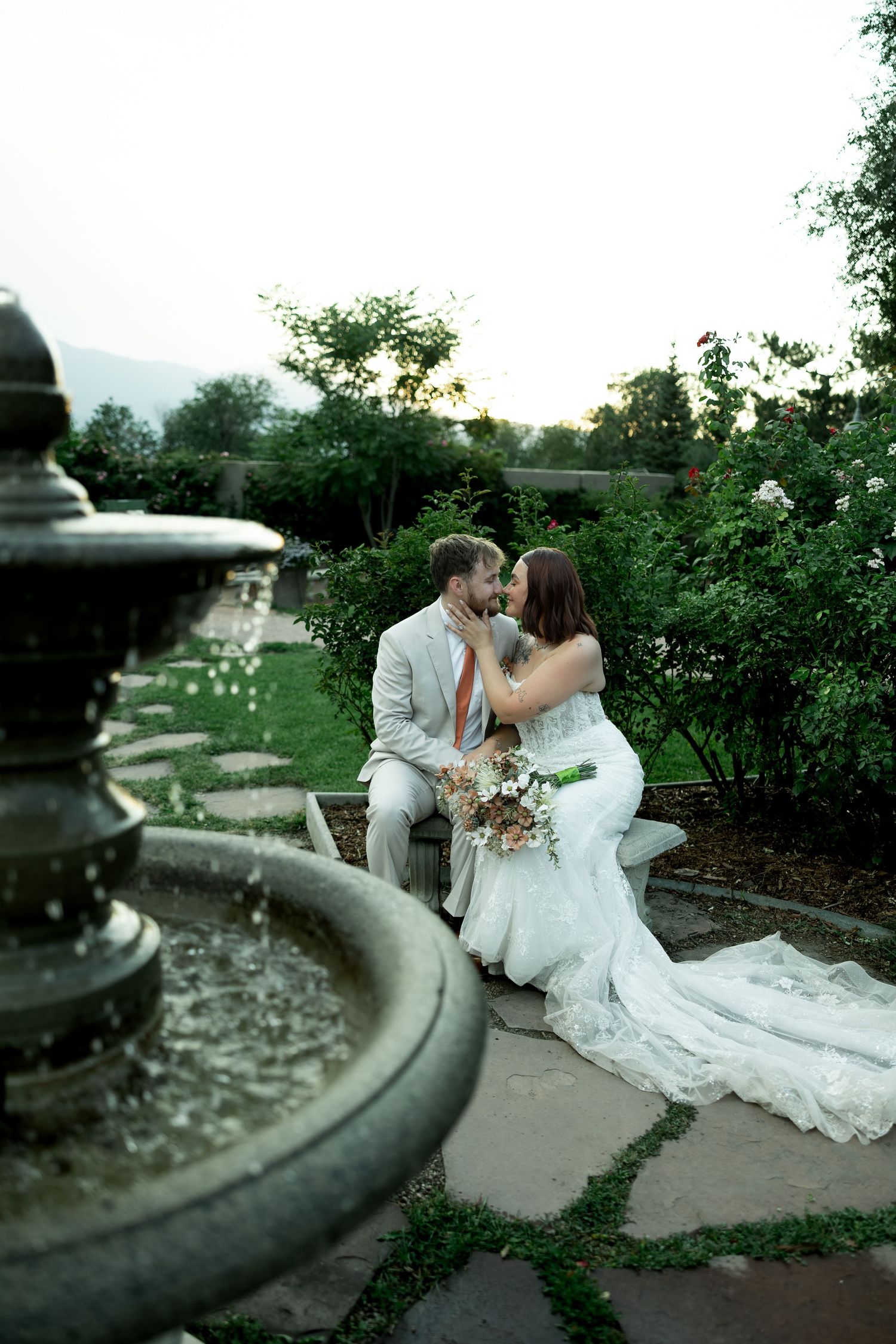 Bride and groom share a romantic kiss on a garden bench near a stone fountain surrounded by lush greenery at sunset.