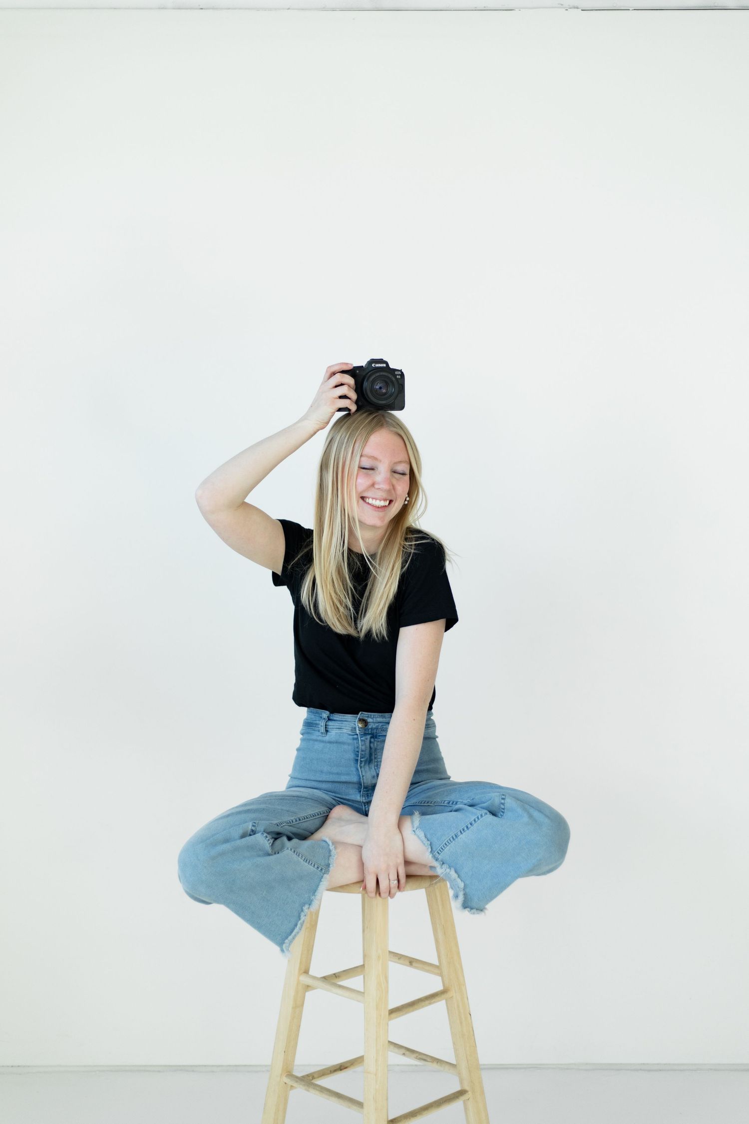 Creative portrait of photographer holding camera while sitting on wooden stool in white studio.