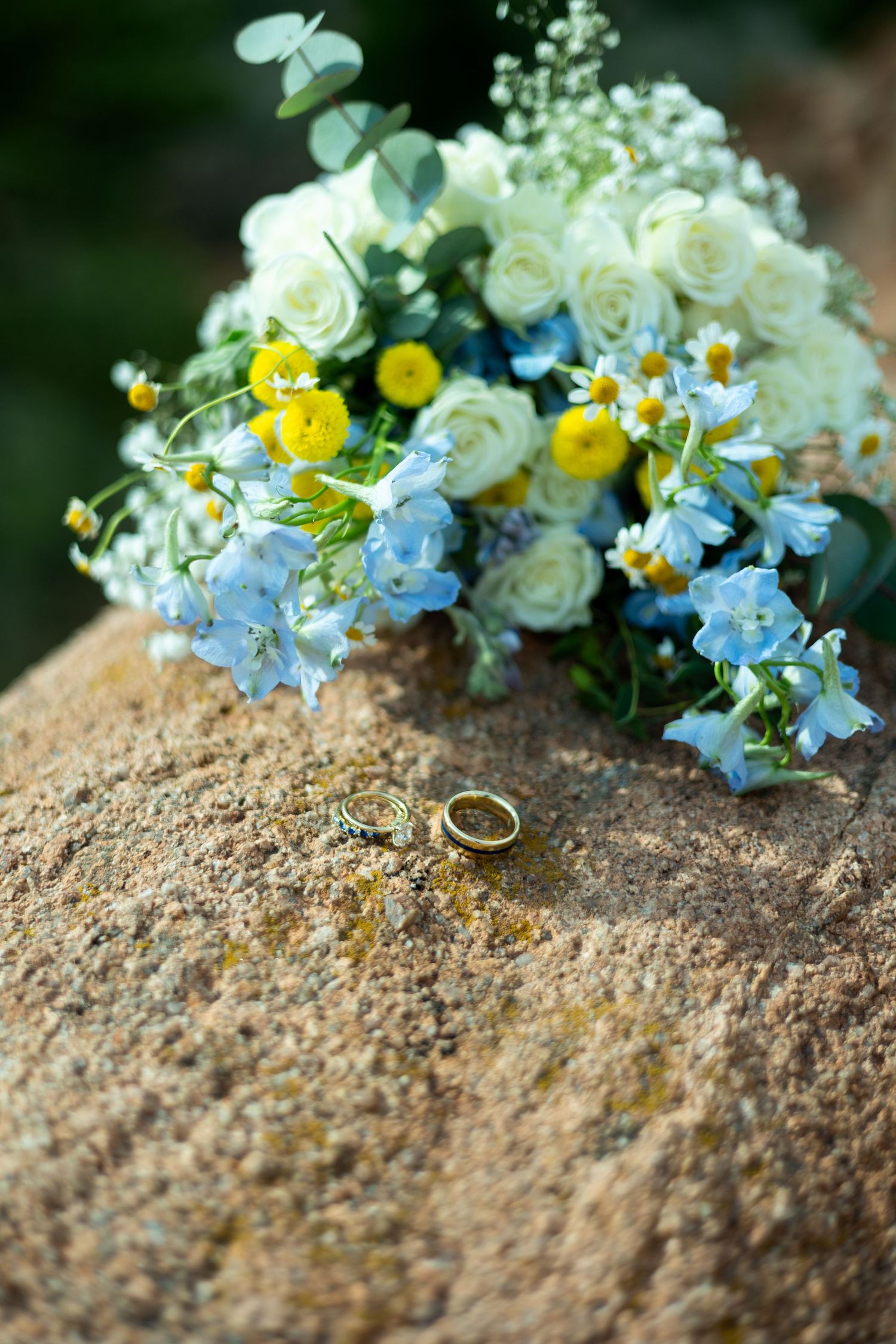Wedding rings nestled among delicate blue and white flowers on a textured stone surface create an elegant bridal detail.
