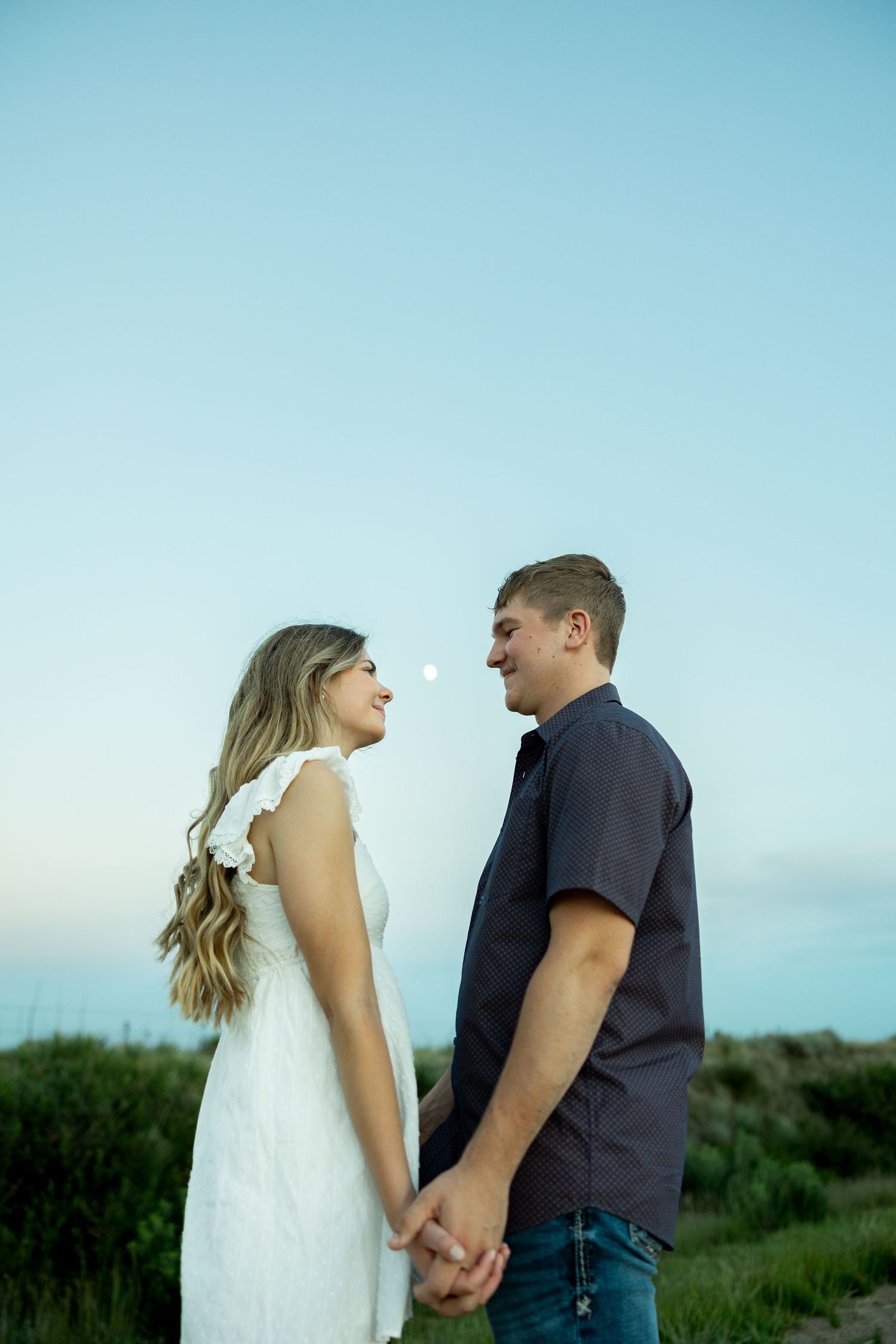 Couple holding hands facing each other in a field at dusk, woman in white dress, man in dark shirt, blue sky background.