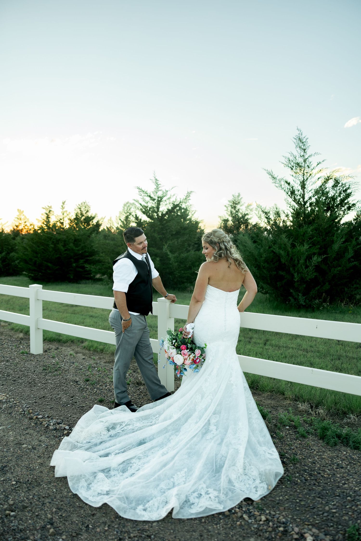 Bride in lace gown with train and groom in vest share a romantic moment by white fence at sunset.