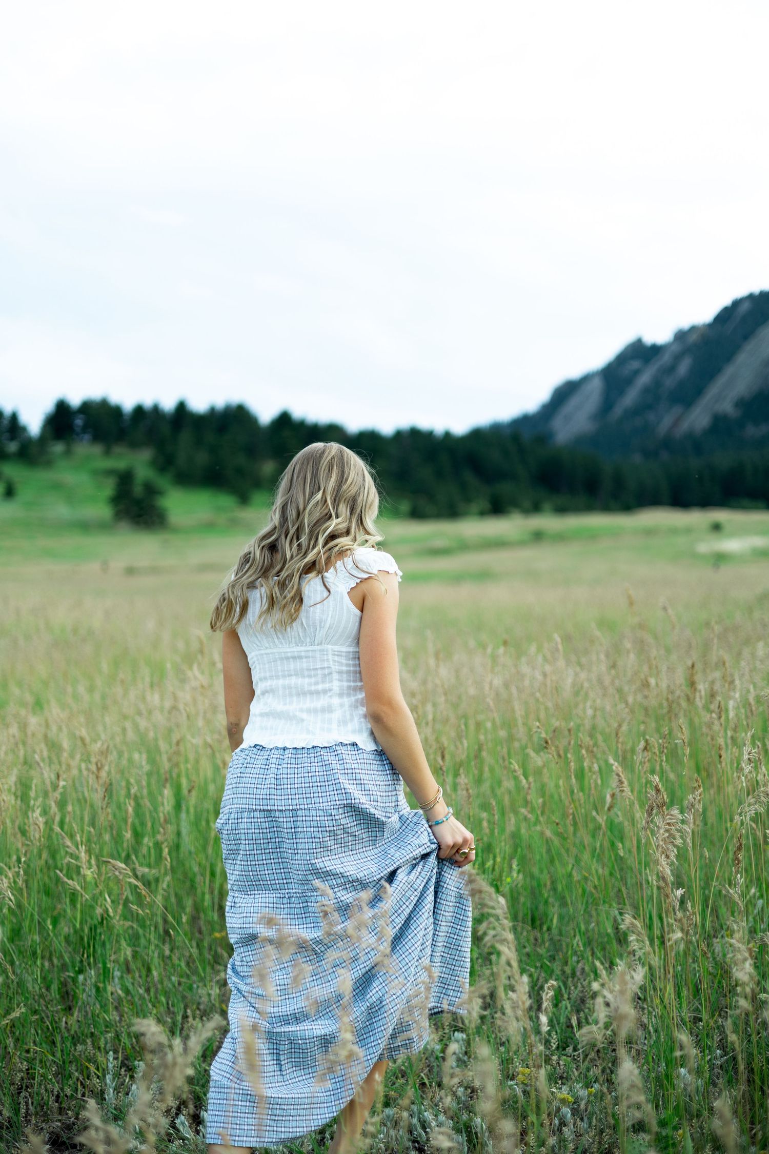 Woman in white top and flowing blue skirt stands in a meadow with mountains in the background at sunset.