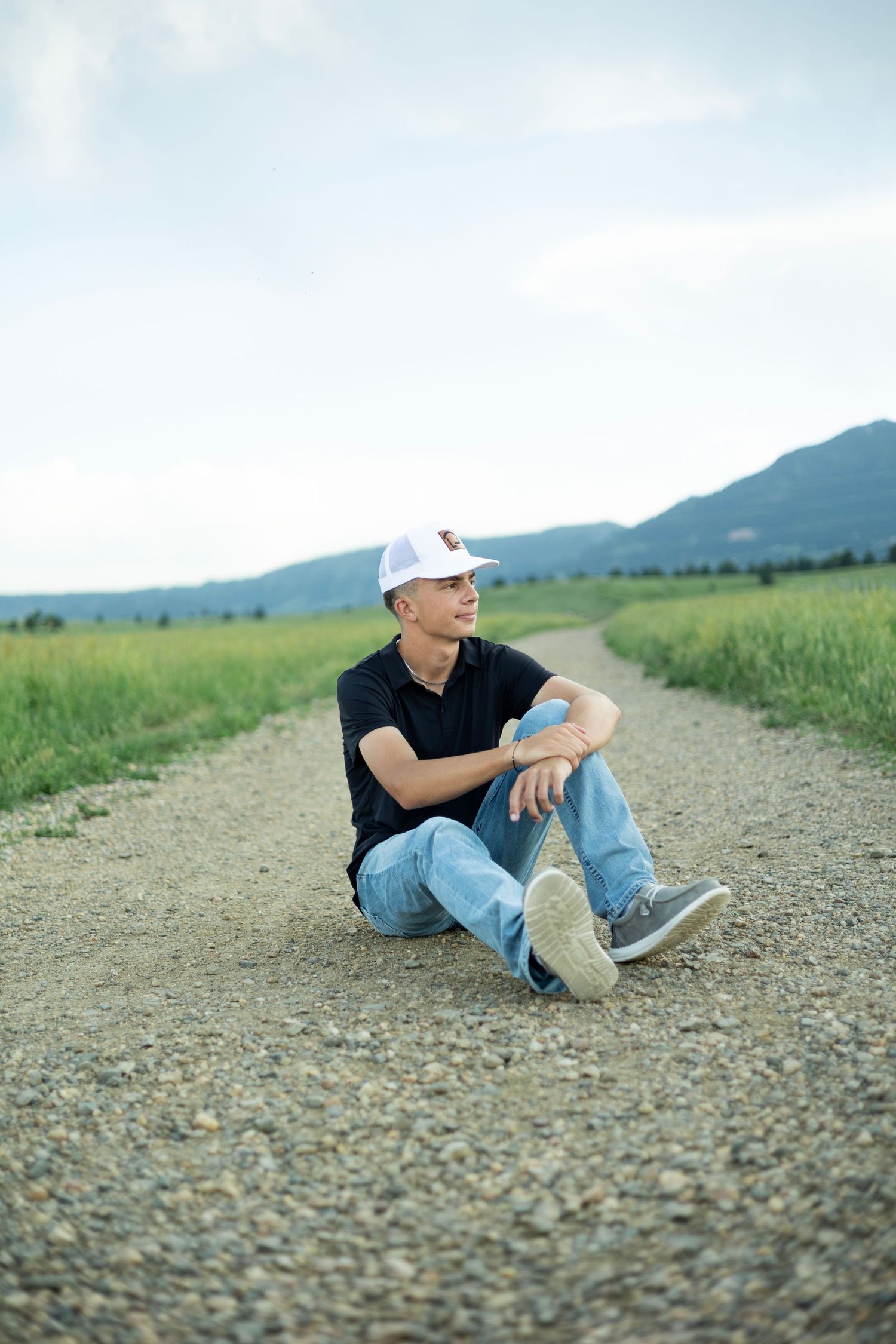 Young man in white cap and jeans sitting on a gravel path surrounded by green fields and mountains.