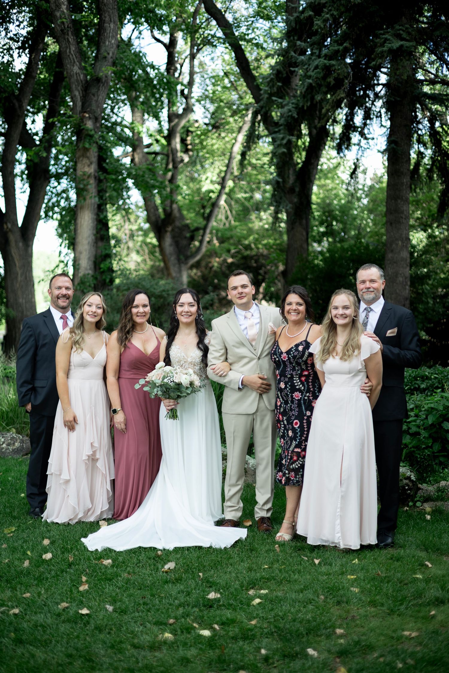Wedding party poses outdoors on green lawn with lush trees, bride in white gown holding bouquet with groom and family.