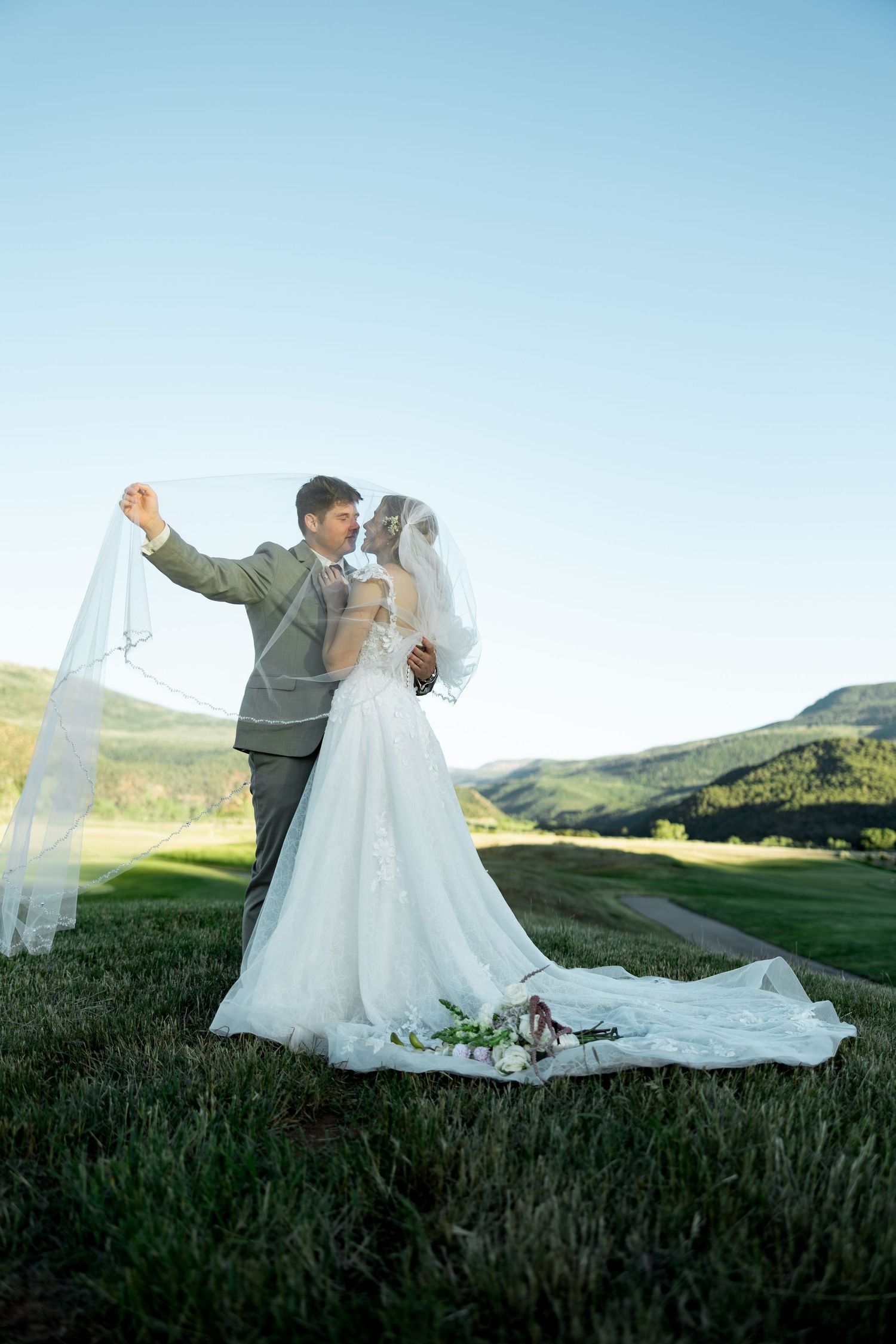 Bride and groom kissing in a green meadow, groom lifting veil, mountains in background on their wedding day.