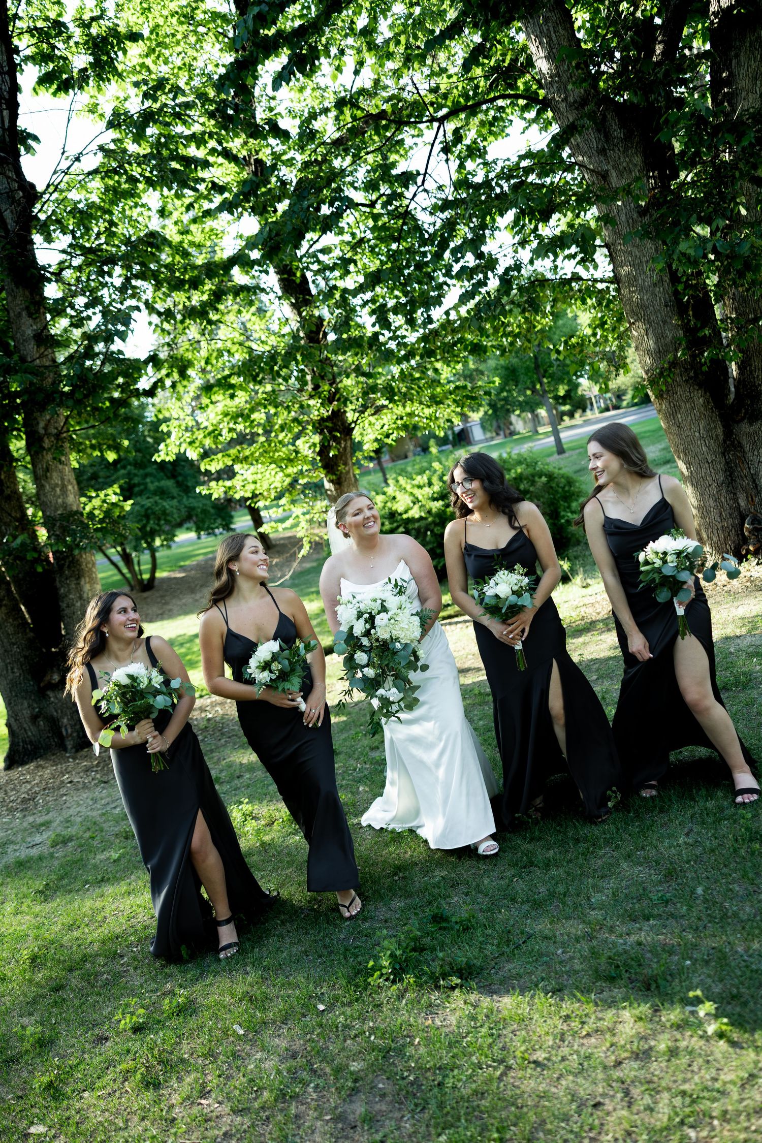 Bride in white gown walks with four bridesmaids in black dresses holding white bouquets outdoors.