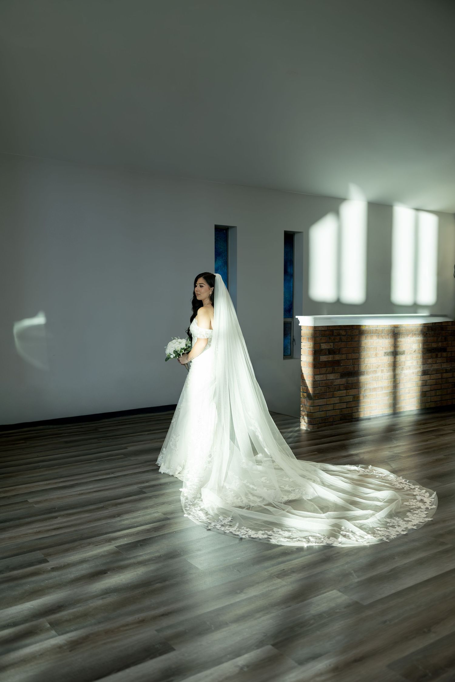 A bride in a flowing white gown with long train poses in an elegant indoor setting with dramatic lighting.