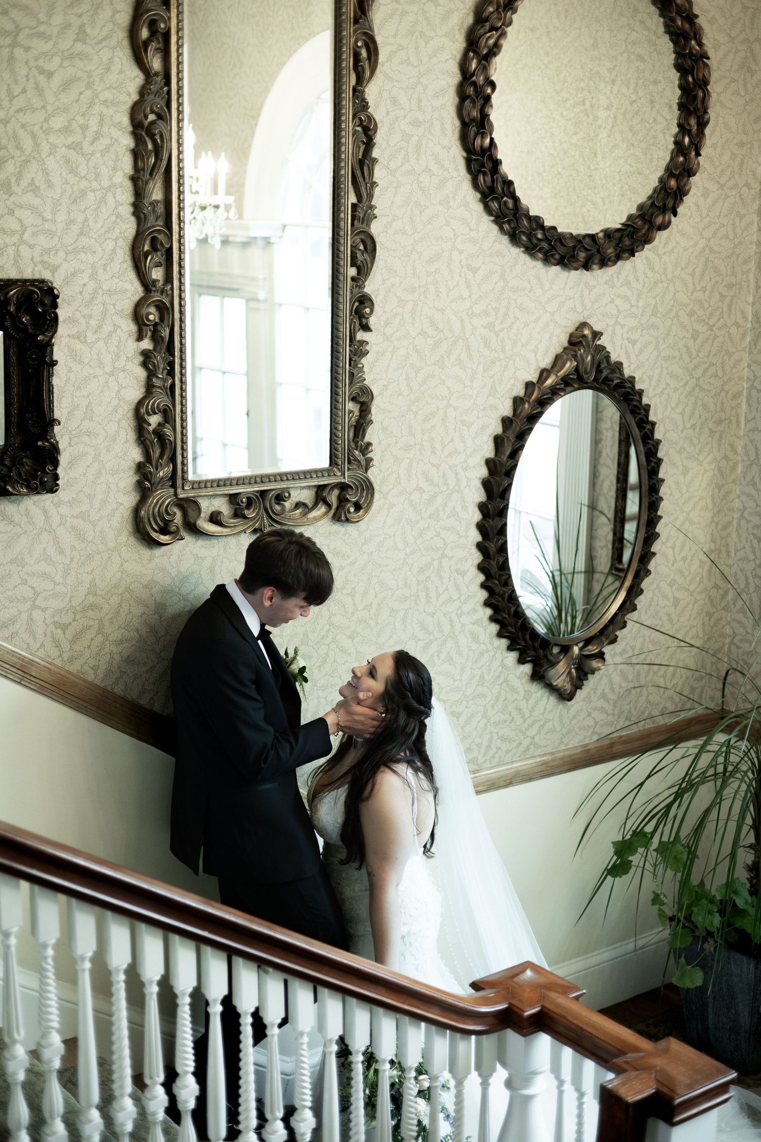 Wedding couple sharing an intimate moment on an elegant staircase with ornate framed mirrors decorating the wallpapered wall.