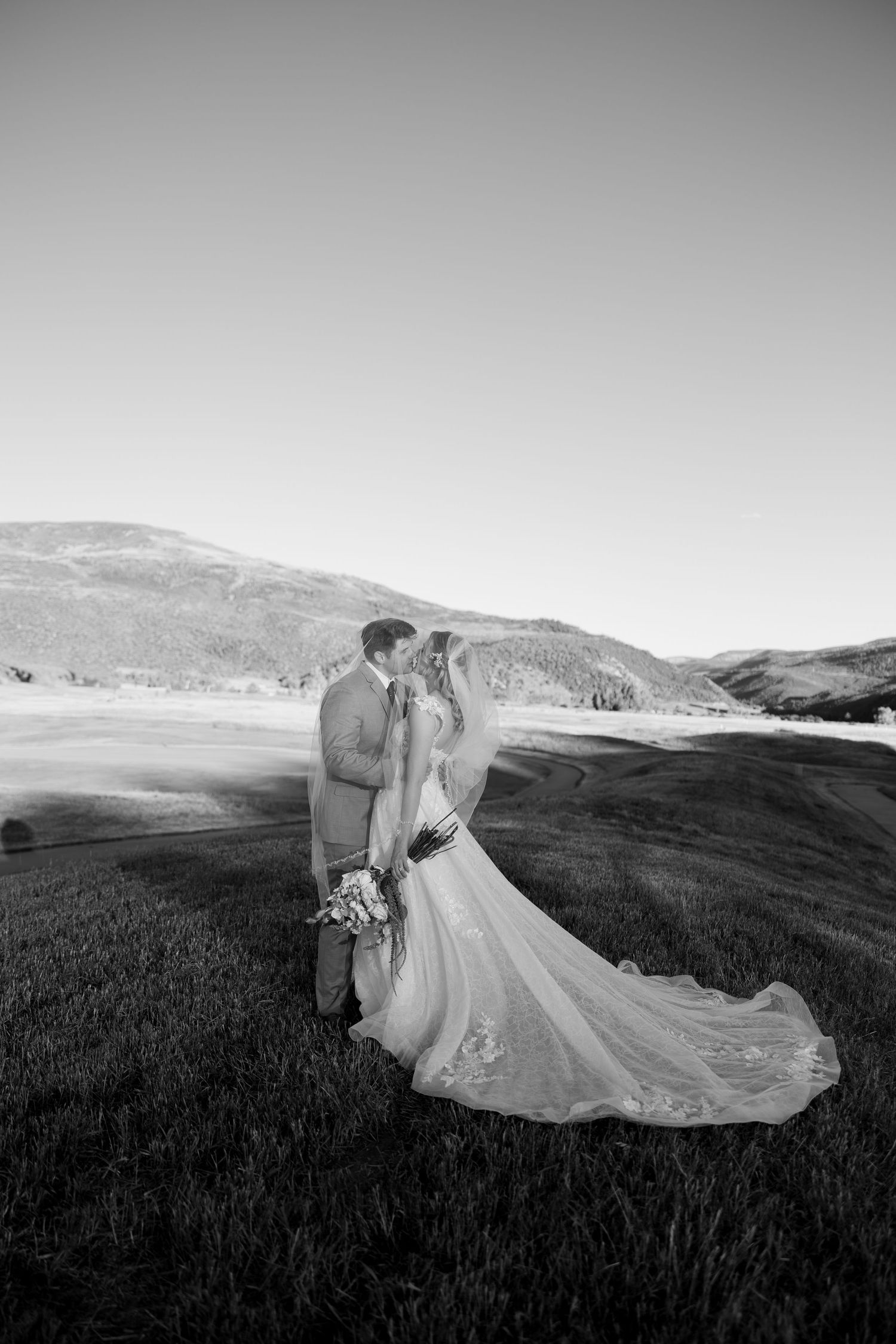 Black and white photo of a bride and groom kissing in a scenic mountain meadow, bride holding bouquet with veil flowing.