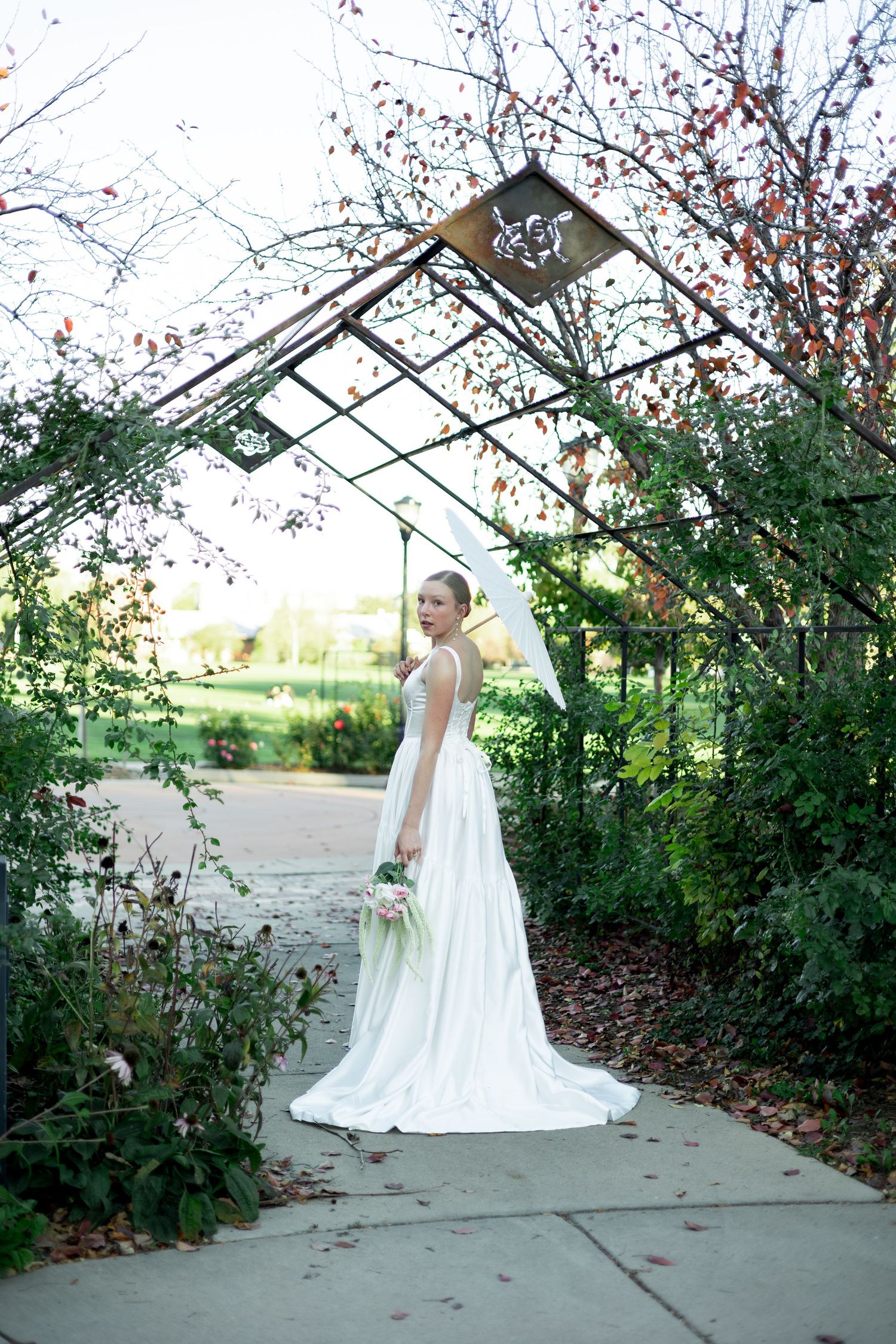 Bride in white gown with veil holding bouquet under garden arbor surrounded by lush greenery.