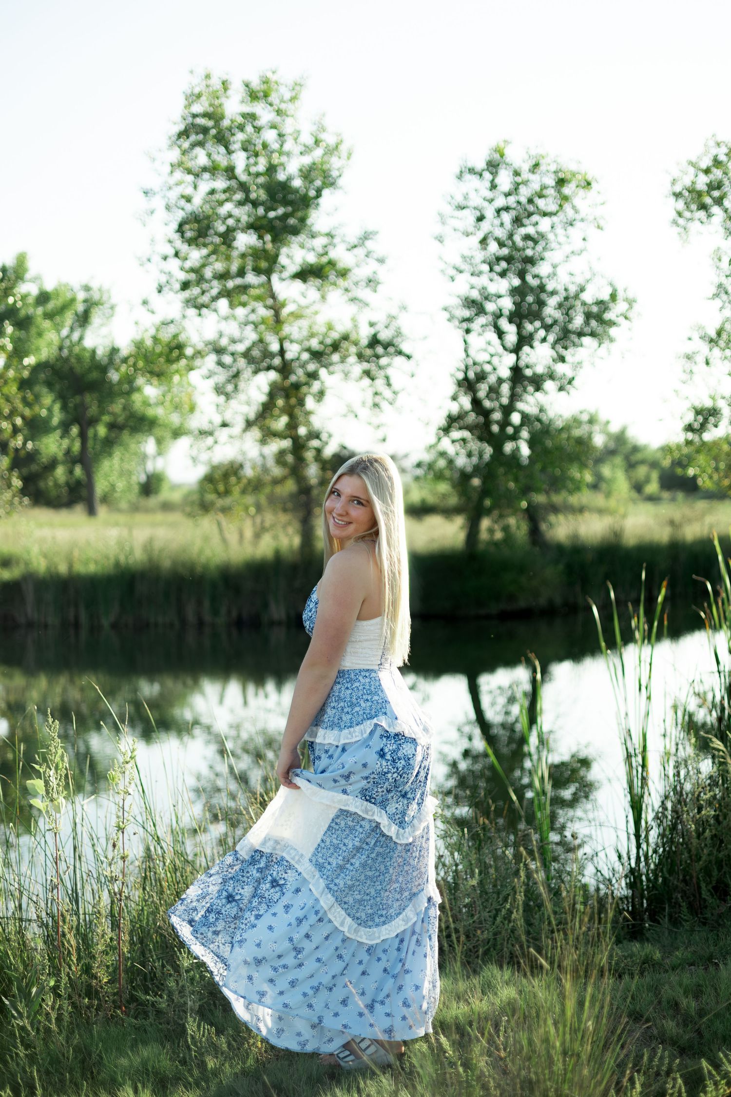 Smiling blonde woman in blue floral maxi skirt standing by a serene pond surrounded by lush green trees.