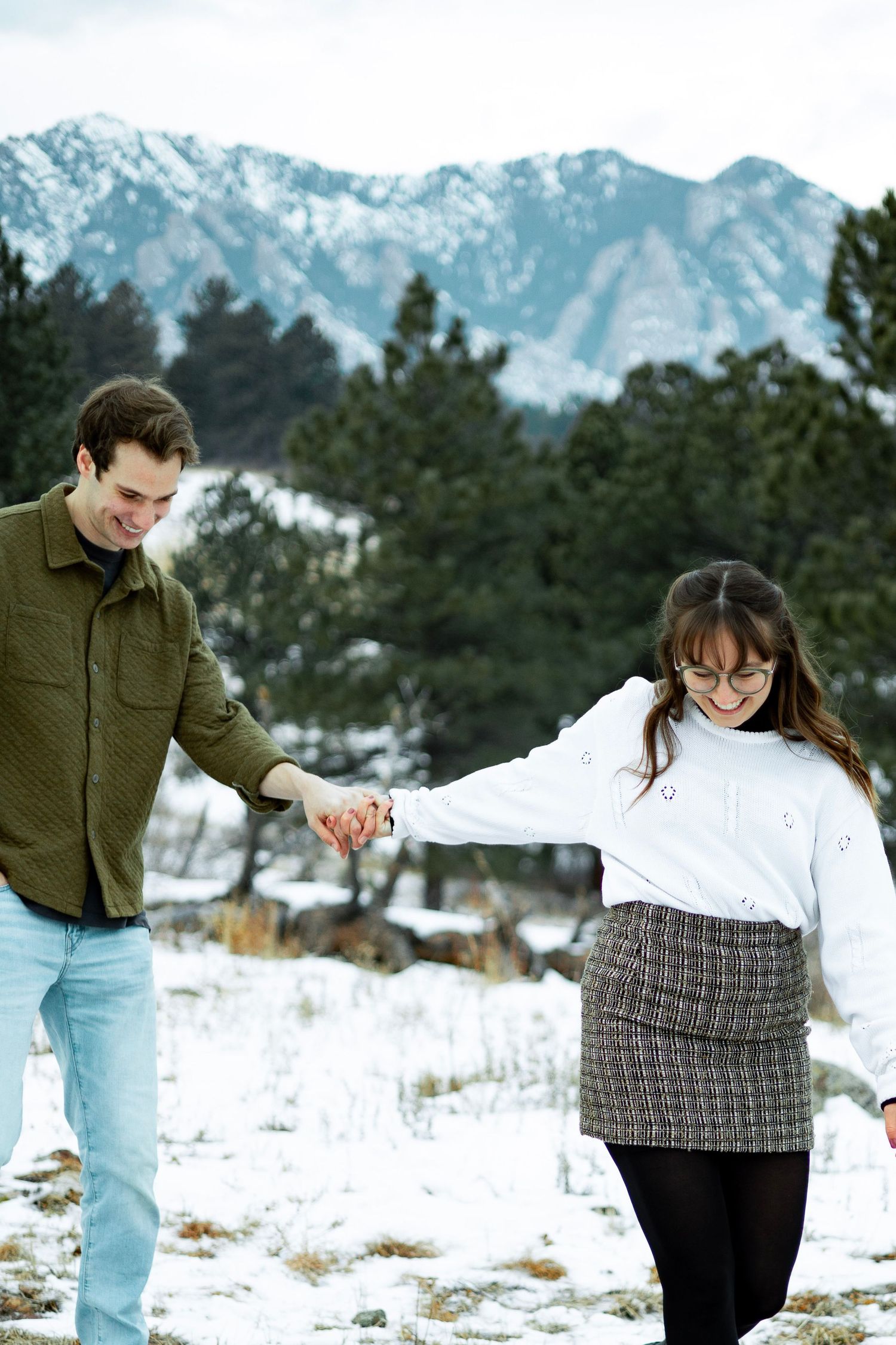 Couple holding hands and walking in snowy mountain landscape, woman in white sweater and plaid skirt, man in green shirt.