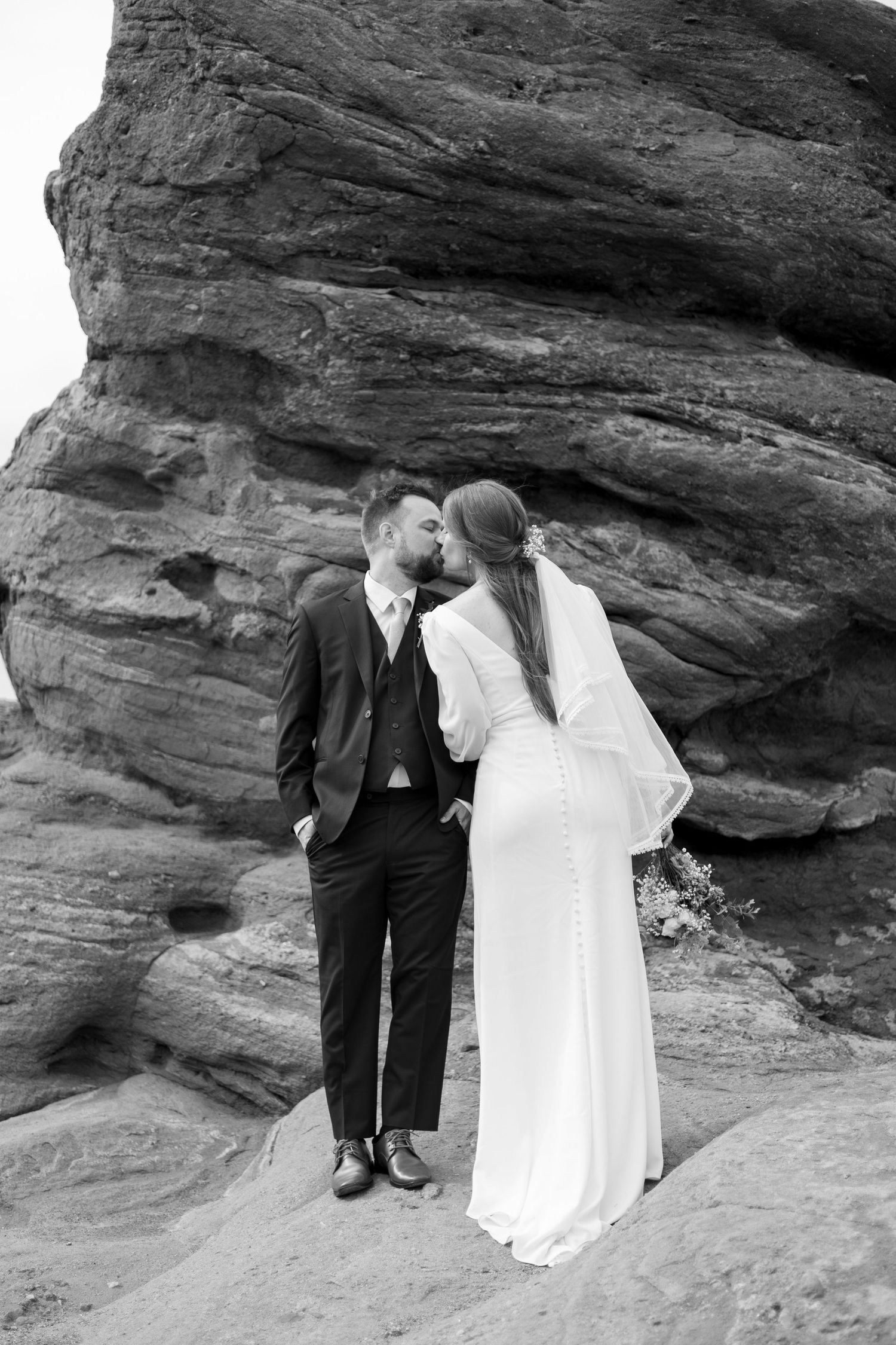 Black and white photo of a bride and groom kissing in front of large rock formations during their wedding day.