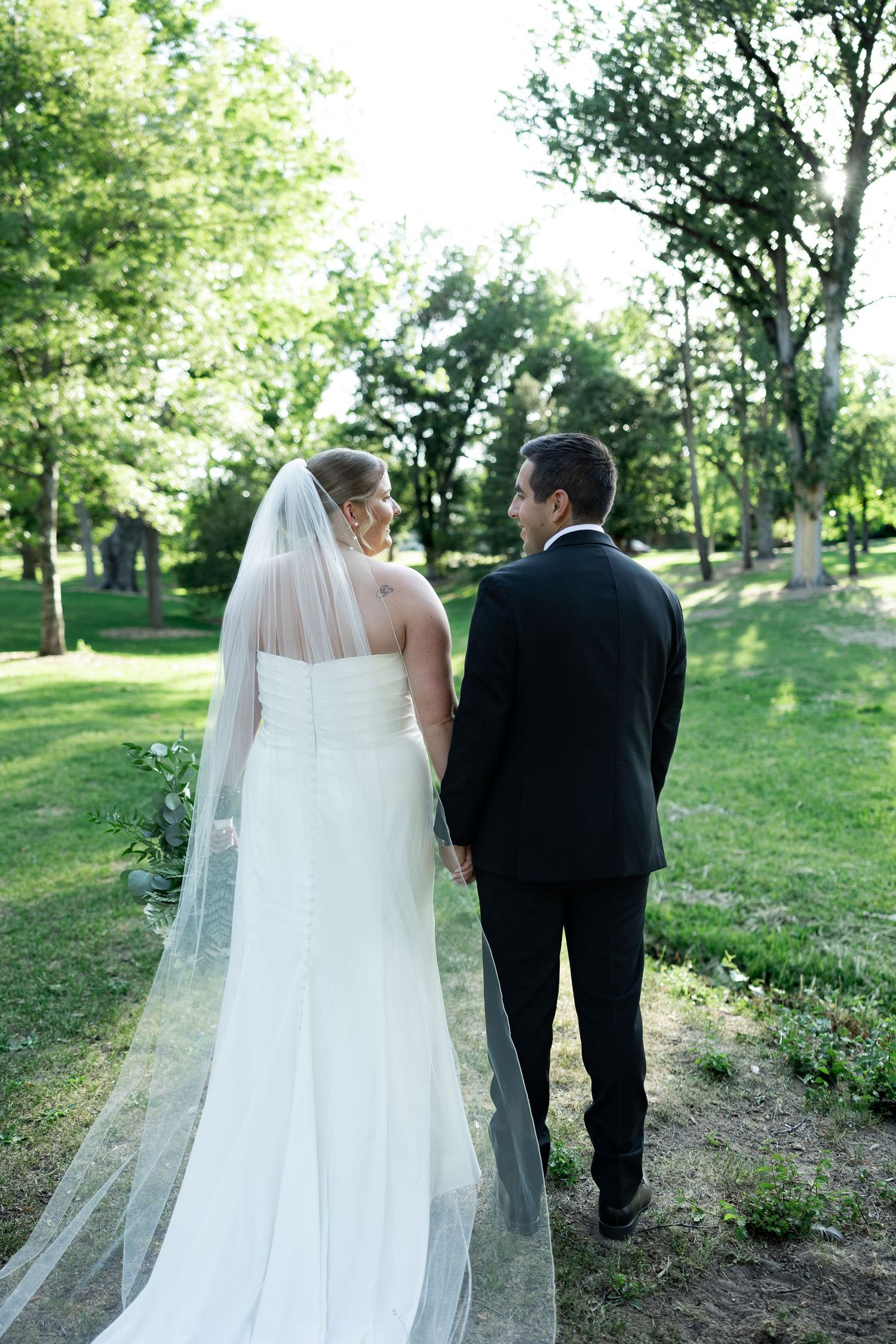 Bride and groom walking hand in hand through a lush green park, viewed from behind on their wedding day.